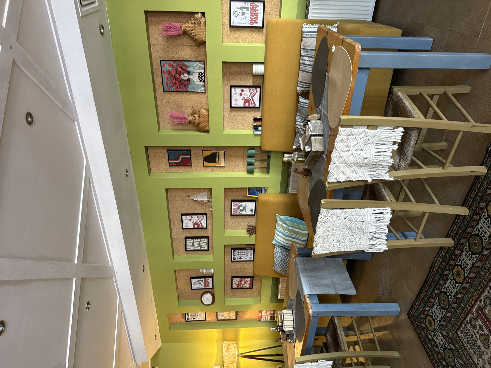 This is a vertical, eye-level, interior shot of a cafe in Tsarevo, Bulgaria, with a distinctly vibrant and eclectic decor. The dominant colors are lime green walls with yellow accents, complemented by natural wood and blue furniture.
The scene showcases a seating area with a long, mustard-yellow booth and several wooden chairs with woven seats, some adorned with macrame throws. The walls are decorated with a series of framed pictures and artisanal woven baskets containing pink dried flowers. A large, patterned rug covers a portion of the floor.
Visible text includes "SIP & SPIN", "OWNER PARTY", "CARAVAN", and "LOVE LIVES". The overall ambiance suggests a lively and artistic establishment, possibly a cafe or a small restaurant, perhaps preparing for or having recently concluded an event. The lighting is bright, indicating it could be daytime, but the exact time of day is not discernible. There are no people visible in the shot, suggesting the space is either empty or the subjects are outside the frame. The setting appears to be a well-decorated indoor space, possibly a commercial establishment, with a focus on visual interest through its unique decorative elements and color palette.
