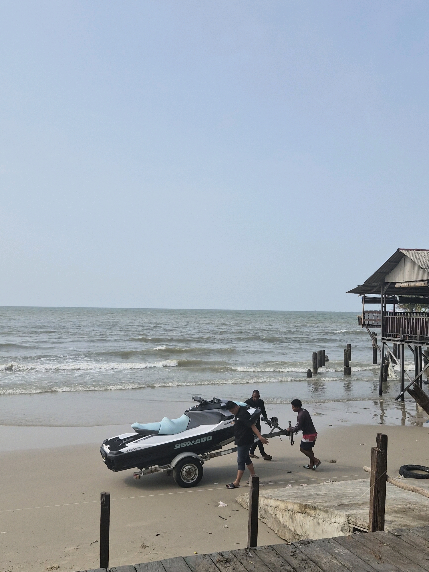 In Balikpapan, Indonesia, three individuals are on a sandy beach near the ocean, preparing to launch a jet ski from a trailer. The jet ski, a black and white Sea-Doo GTX model, is positioned on a trailer with a single wheel visible. Two men are actively pushing the trailer forward, while a third man stands behind them. The ocean exhibits moderate waves, and a weathered, wooden stilt house is situated on the right side of the frame, extending over the water. The sky is clear and pale blue. The foreground consists of a sandy beach and wooden planks, suggesting a pier or boardwalk.