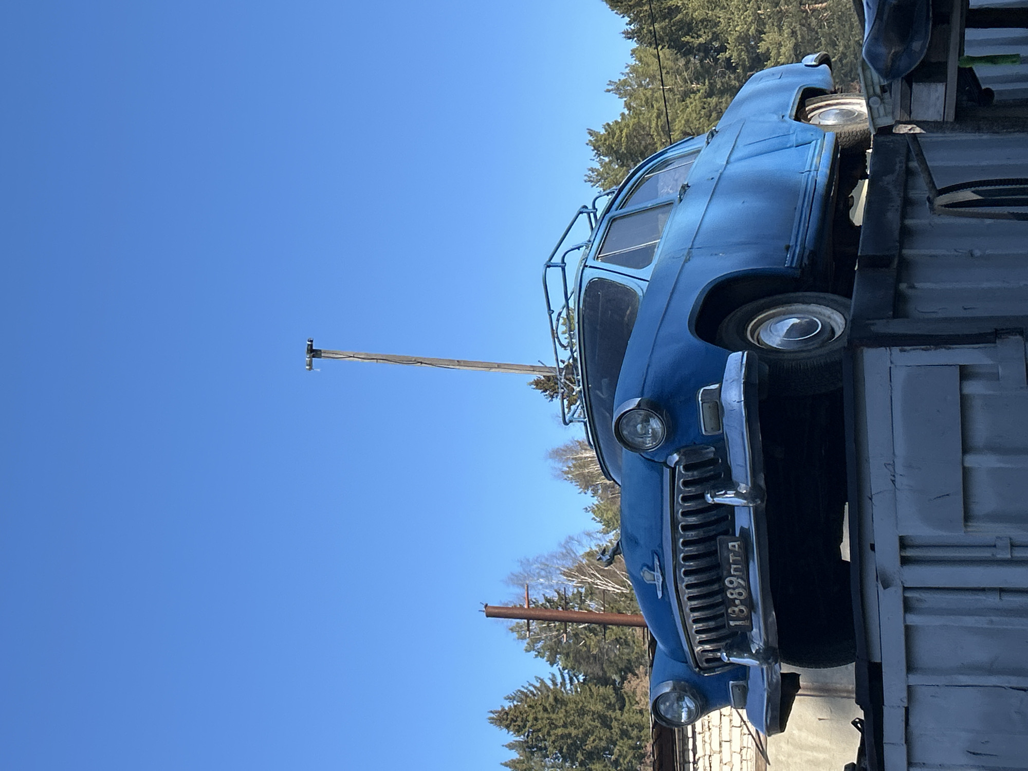A blue vintage car with the license plate "18-89 UTA" is mounted on a white metal structure. The car has a roof rack. Telephone poles are visible in the background against a clear blue sky and a line of trees. The location is Chaikovsky, Russia.