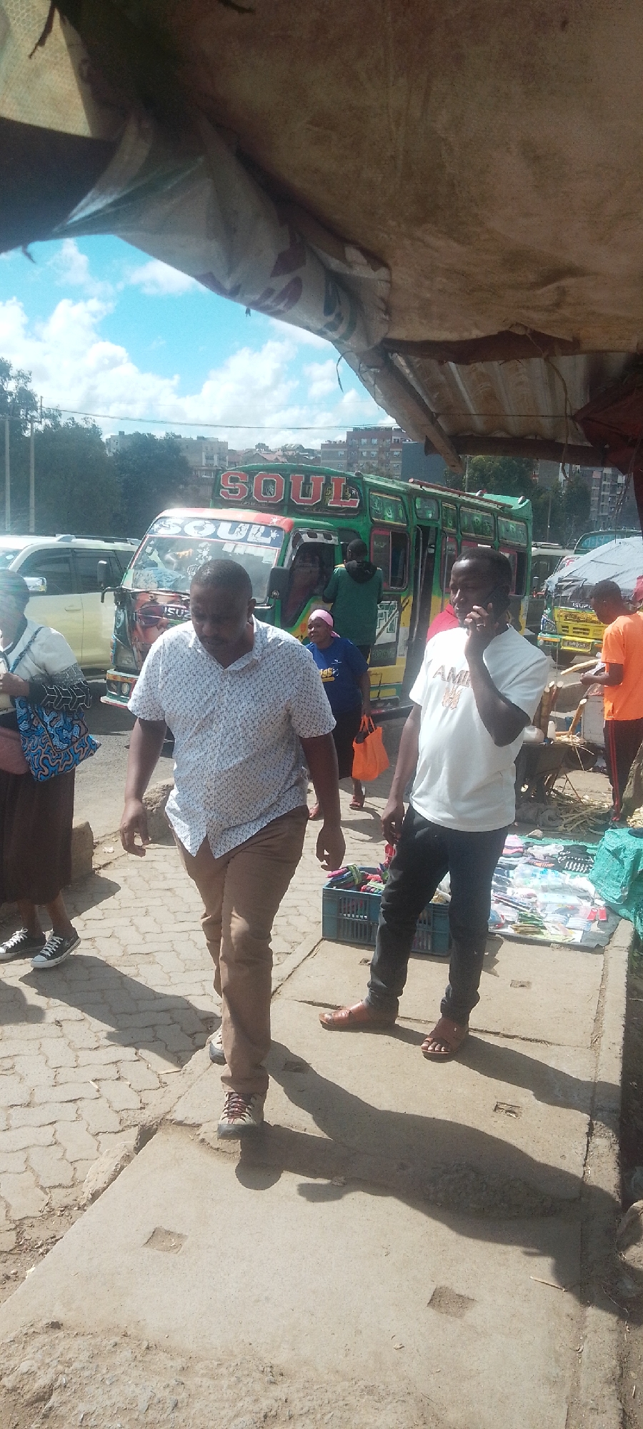 This image depicts a bustling street scene in Nairobi, Kenya, under strong daylight with a blue sky and scattered white clouds. An overhead, makeshift canopy of weathered fabric and corrugated metal partially shades the foreground, casting distinct shadows on the ground.
The central feature is a highly decorated green, yellow, and red Isuzu matatu (public transport bus), emblazoned with "SOUL" on its roof and front. Several individuals are present around and near the matatu, indicating active transit and street commerce.
In the immediate foreground, a male subject walks towards the lower left, wearing a white patterned short-sleeved shirt, tan trousers, and light-colored sneakers. To his right, another male subject stands, holding a black mobile phone to his right ear. He wears a white short-sleeved t-shirt with a dark "AMI" graphic and dark trousers, paired with brown sandals. Adjacent to him, a blue plastic crate filled with colorful merchandise and other items for sale are displayed on the ground.
To the left of the walking man, a partially visible female subject in a patterned white top and dark skirt carries a blue patterned bag. Near the matatu's open passenger door, a male subject in a dark green hoodie is visible, and a female subject in a pink headscarf and blue top holds an orange bag. To the far right, a male subject in an orange t-shirt stands near a small stall with bundled goods. The ground consists of concrete slabs transitioning to interlocking pavers. In the background, other vehicles, trees, and urban buildings on a hillside are visible.