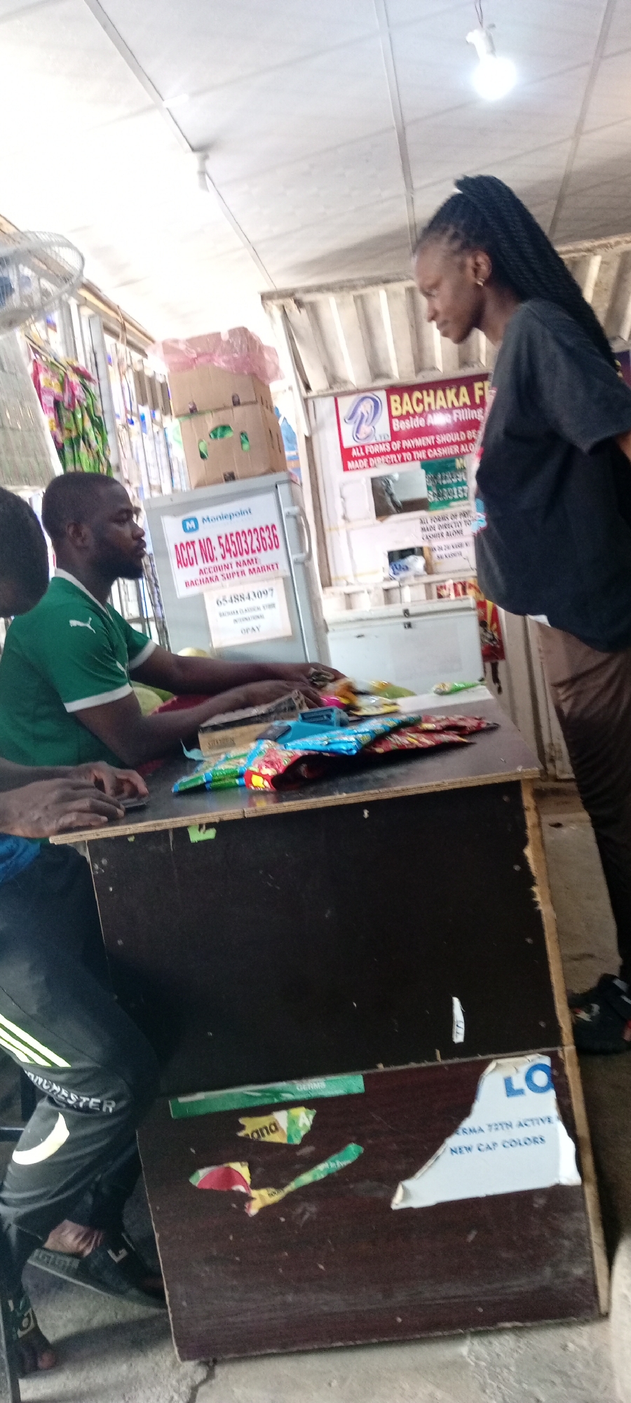 This media file captures a scene inside a small shop or market stall, likely in Aviation Quarters, Nigeria. The primary subjects are three individuals. A man wearing a green shirt with white trim is seated behind a counter, appearing to be working with some items. To his left, another person is partially visible, also seated. Standing opposite the man at the counter is a woman with braided hair, looking down.
The setting is indoors, characterized by a white, tiled ceiling with visible fluorescent lighting and what appears to be a ceiling fan. The counter itself is cluttered with various packaged goods, possibly snacks or small items, suggesting a retail environment. Behind the counter, there are signs and a refrigerator. One prominent sign displays "Moniepoint" with an account number and "BACHAKA SUPER MARKET" as the account name, indicating a financial transaction point or a business name. Another sign reads "BACHAKA FC" and "Beside Almo Filling," providing further location context.
The activity appears to be a transaction or customer service interaction at the counter. The man's hands are on or near the counter, possibly handling items or a device. The woman is looking down, perhaps at the counter or items, and her posture suggests engagement in the interaction.
Visible text includes brand names like "Moniepoint" and "OPAY," business names like "BACHAKA SUPER MARKET," and informational text on signs related to payment procedures ("ALL FORMS OF PAYMENT SHOULD BE MADE DIRECTLY TO THE CASHIER ALONE"). There are also partially visible labels on packaged goods and some shredded pieces of paper or stickers on the counter and the counter's facade. The overall impression is of an active, everyday commercial scene.
