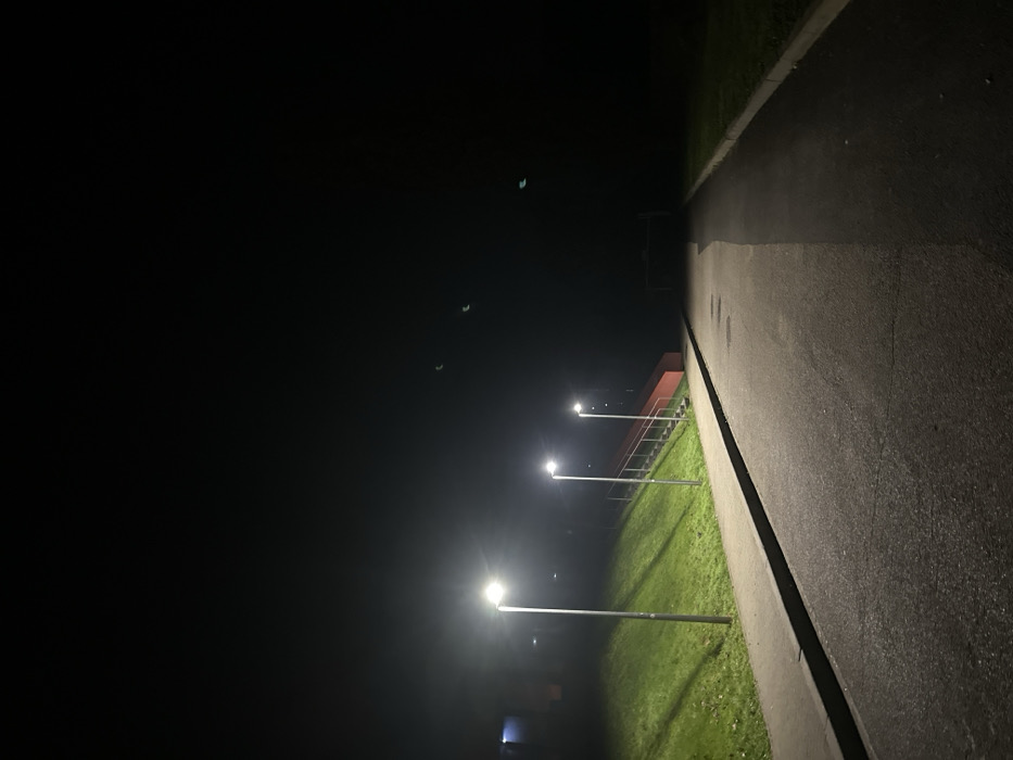 A dark, nighttime scene shows a paved pathway on the right, bordered by a strip of grass illuminated by three evenly spaced streetlights. The ground slopes down toward the left. Behind the streetlights, a red building with metal railings is partially visible. The sky is dark with only a few small, faint light spots. The overall perspective is slightly angled, giving a sense of depth to the scene.