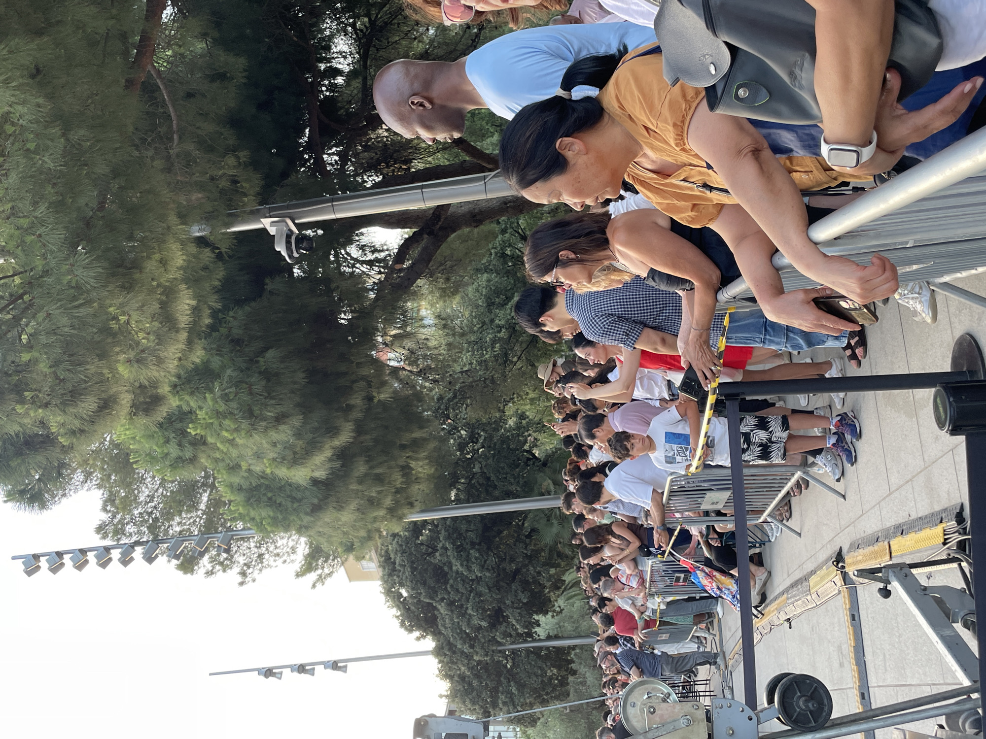 The image shows a crowd of people gathered outdoors, with many standing behind a metal barrier. The environment suggests a public space with trees and lighting poles visible. In the foreground, a young boy in a t-shirt and shorts stands near the barrier. To his right, a woman in an orange top rests her arms on the barrier, and a black bag hangs from her shoulder. Behind her, a man with a shaved head wearing a light blue t-shirt looks to the side. The crowd is diverse in age and ethnicity. Several lighting poles with multiple lights are visible in the background, along with dense green trees, suggesting an urban park or outdoor event venue. The presence of barriers implies a controlled viewing area, possibly for a performance or gathering. The lighting suggests it is daytime, likely late afternoon or early evening.