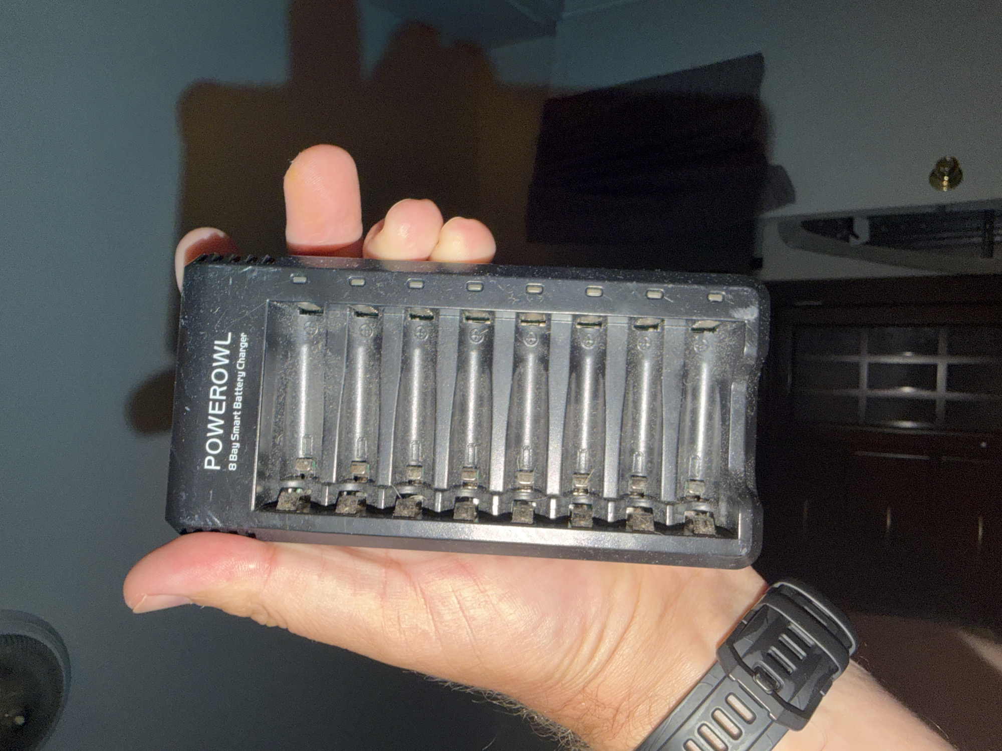 A hand in Pimmit Hills, United States of America, holds a black Powerowl 8-Bay Smart Battery Charger with multiple AA or AAA batteries inserted. The charger is mostly full, with batteries occupying most of the slots. The hand is positioned on the left side of the image, supporting the charger. The background is dark and out of focus, suggesting an indoor, dimly lit environment. The charger appears to be made of plastic and shows some signs of dust or wear in the battery slots. A watch is visible on the wrist of the hand holding the charger.
