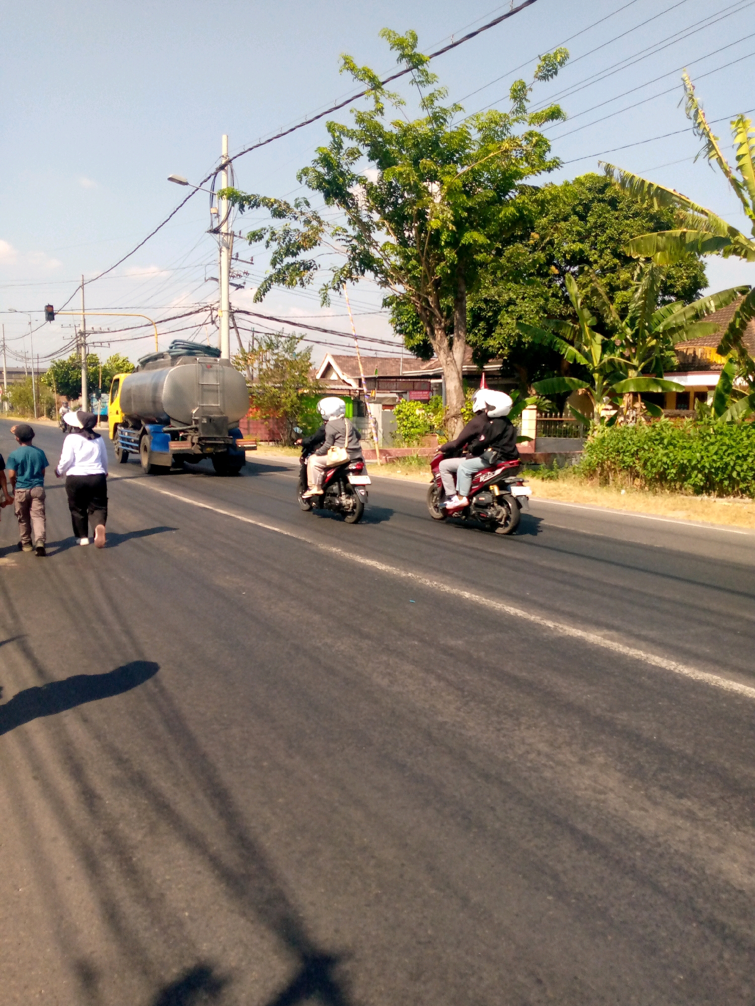 On a sunny day in Sumberwono, Indonesia, a yellow truck carrying a large cylindrical tank is driving down a paved road. The truck has a blue chassis and is partially obscured by a large tree on the right side of the frame. Two people are walking on the side of the road, with the person closer to the truck wearing a white shirt and black pants, and the person further away wearing a blue shirt and grey pants. A motorcycle with two occupants, both wearing helmets and white shirts, is passing the truck on the right side of the road. The environment is rural, with trees and houses visible in the background. The road has a white line marking the lane and shadows of power lines are cast across the asphalt.