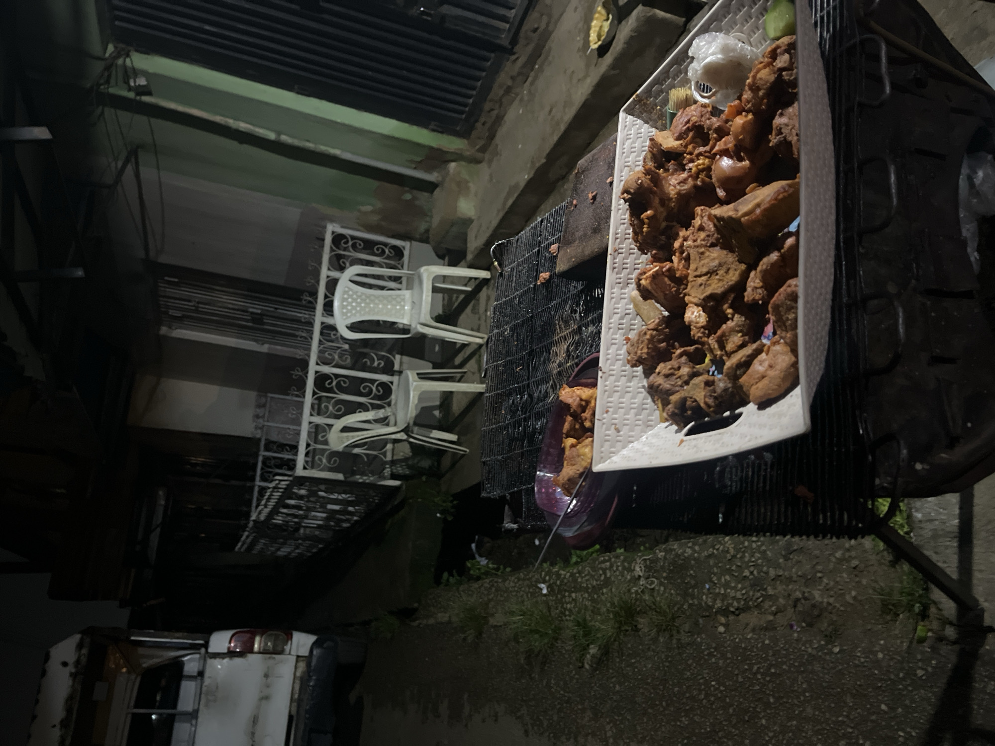 An outdoor scene at night shows a food stall with several containers of cooked meat, possibly fried chicken or goat meat, arranged on a grill. A white plastic chair and a metal fence are visible in the background. The stall appears to be set up along a roadside in Jos, Nigeria, as indicated by the presence of a white truck parked to the left of the frame. The lighting is dim, suggesting it might be late evening or early morning. The overall atmosphere is that of a local eatery, possibly a street food vendor preparing for or serving customers.