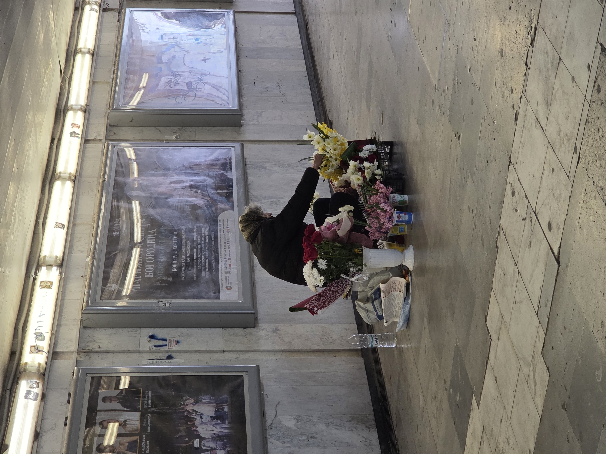 A middle-aged person is kneeling on a gray tiled floor in what appears to be an underpass or hallway, potentially in Varna, Bulgaria. The person is surrounded by an array of flowers, including daffodils, roses, and carnations. A black plastic crate and several containers hold the bouquets. A bottle of water and crumpled paper are next to the flowers. Behind the person, the wall is lined with rectangular frames containing posters. The posters promote events, possibly at a local theater or cultural center, including a performance entitled "Bogorodica." A row of fluorescent light fixtures runs along the top of the wall.