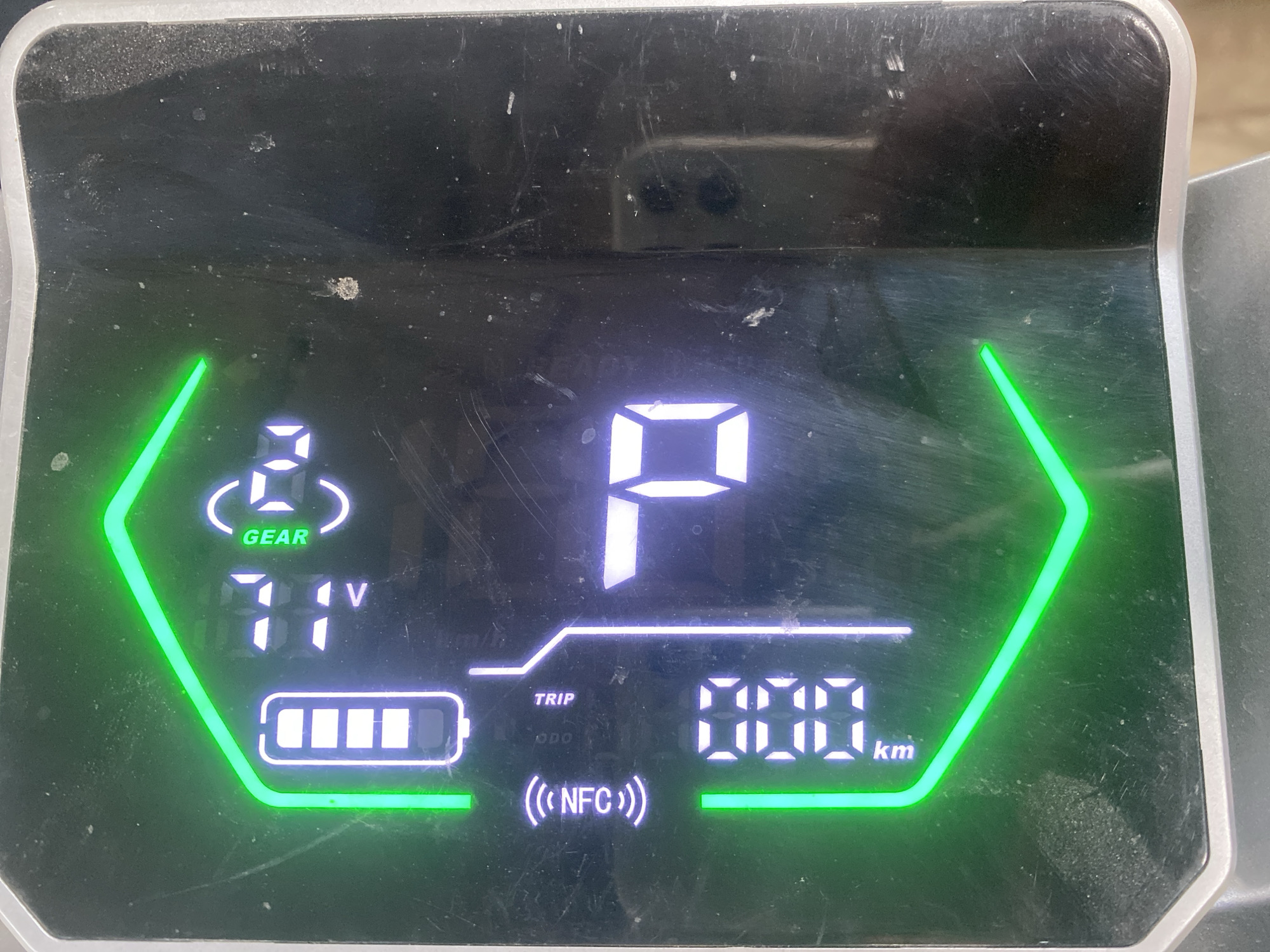 An electronic display shows battery charge as a vertical bar, an NFC icon, a mileage reading of "000 km," and a gear indicator with the text "GEAR" and a stylized wheel. There are also digital readouts of "31V" and "6." The display is dirty and scratched. The surrounding environment is out of focus, but appears to be a dark, possibly outdoor setting. The image was taken in Balta, Ukraine.