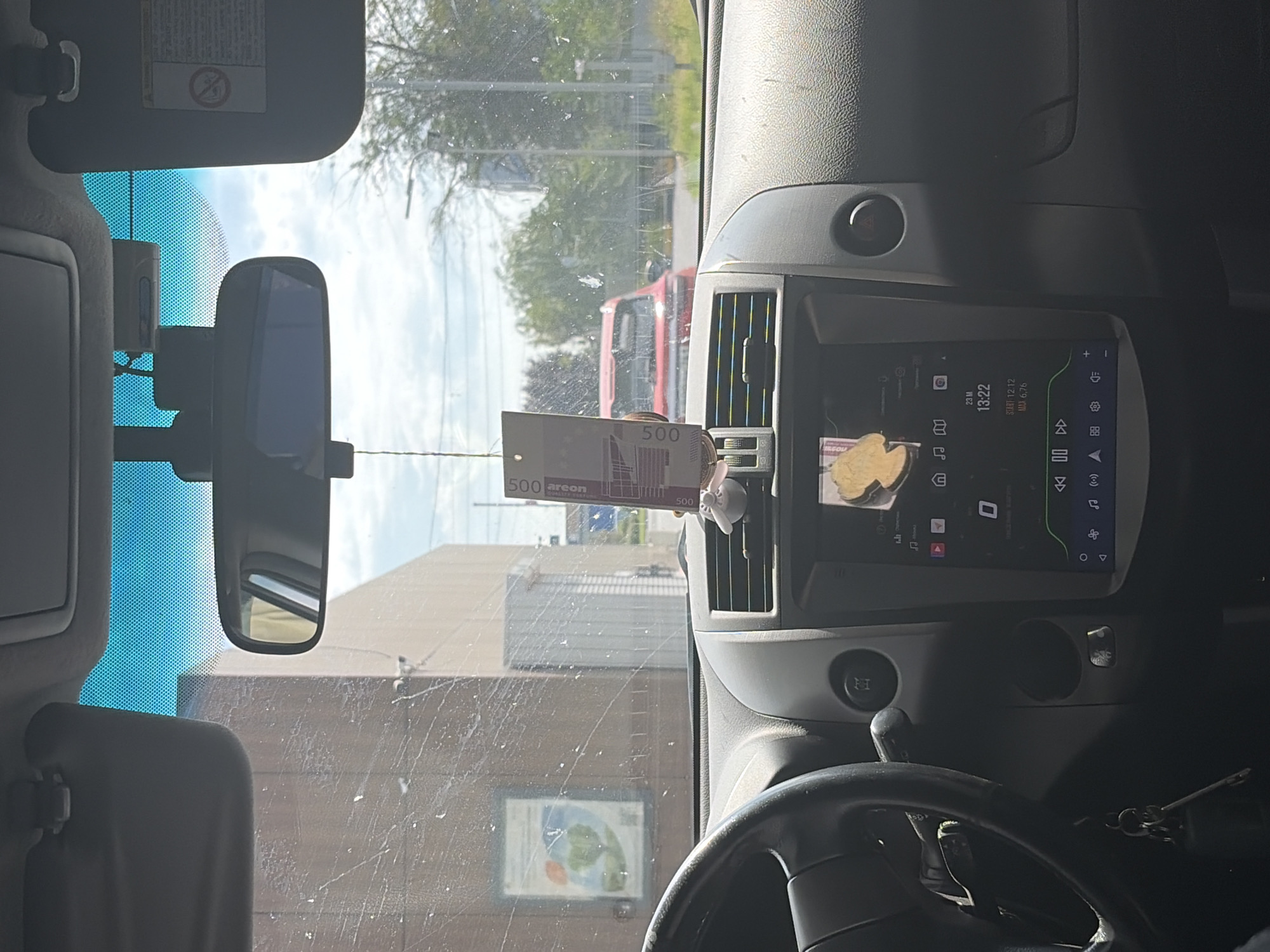 The image provides an interior view of a car, looking forward through the windshield. The dashboard is visible, with a steering wheel on the left and a central infotainment screen displaying a clock at 13:22 and what appears to be a route navigation or music application. A novelty air freshener is hanging from the rearview mirror, along with a string featuring what looks like a Euro banknote. Outside the car, through the somewhat smudged windshield, a red car is visible in the distance, along with trees and a building with signage. The sky is bright and blue, suggesting daytime. The presence of the Euro banknote, a currency not used in Russia, combined with the visible building and surrounding environment, suggests the photo was taken at a location that might be confused for Rostov-on-Don, Russia due to similar building architecture and vegetation. However, without more conclusive evidence, it is difficult to definitively place the location.