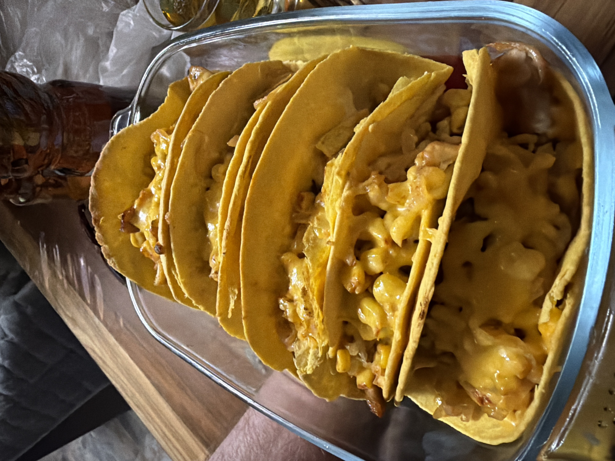 A close-up, high-angle shot shows a glass baking dish filled with several corn tortillas folded and filled with cheese and a corn and chicken mixture. The tortillas are golden yellow and appear to be baked until crispy. The cheese is melted and gooey, covering the filling. The dish is placed on a wooden surface. In the background, out of focus, is a dark brown jug and a glass of amber liquid. The lighting is warm and ambient. The scene is likely set indoors, possibly in a home kitchen or dining area. There is no discernible indication of the city of Низинка Снт, Russia.