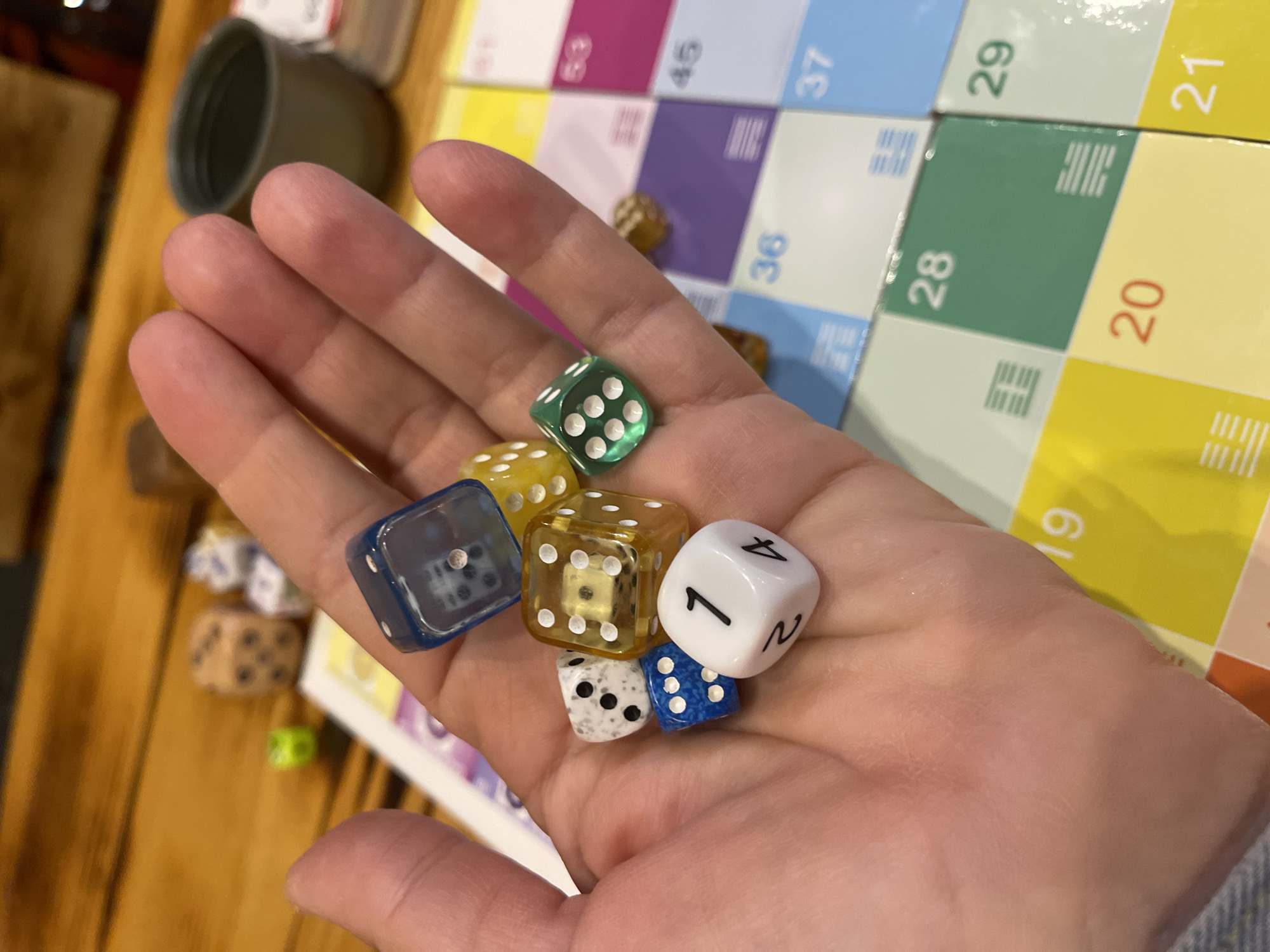 A close-up image captures a person's left hand holding a collection of dice over a colorful board game. The hand, with fair skin, is open and elevated, presenting six dice. These dice include: one white d6 showing the number '1' on its top face, with '2' and '4' visible on adjacent sides; one small blue d6 with white pips showing '5'; one small mottled white and grey d6 with black pips showing '5'; one larger translucent yellow d6 with white pips showing '5'; one larger translucent blue d6 with white pips showing '1'; and one larger translucent green d6 with white pips showing '6'.
Beneath the hand and extending into the background, a board game composed of square tiles is visible. The tiles feature a variety of colors including yellow, light blue, green, pink, and purple. Several tiles display numbers, such as '19', '20' (in red), '21', '28' (in green), '29' (in green), '36' (in blue), '37' (in blue), '45', and '53' (in pink). Many tiles also exhibit symbols resembling I Ching hexagrams. The board has a glossy finish.
In the background, on the wooden table supporting the game, additional dice are scattered: a light brown or wooden die showing '4' pips, a small bright green die, and another small white die with blue markings. A dark grey or olive-colored mug is visible in the upper right background. The image contains no discernible visual information identifying a specific location within the city of Kaliningrad, Russia.
