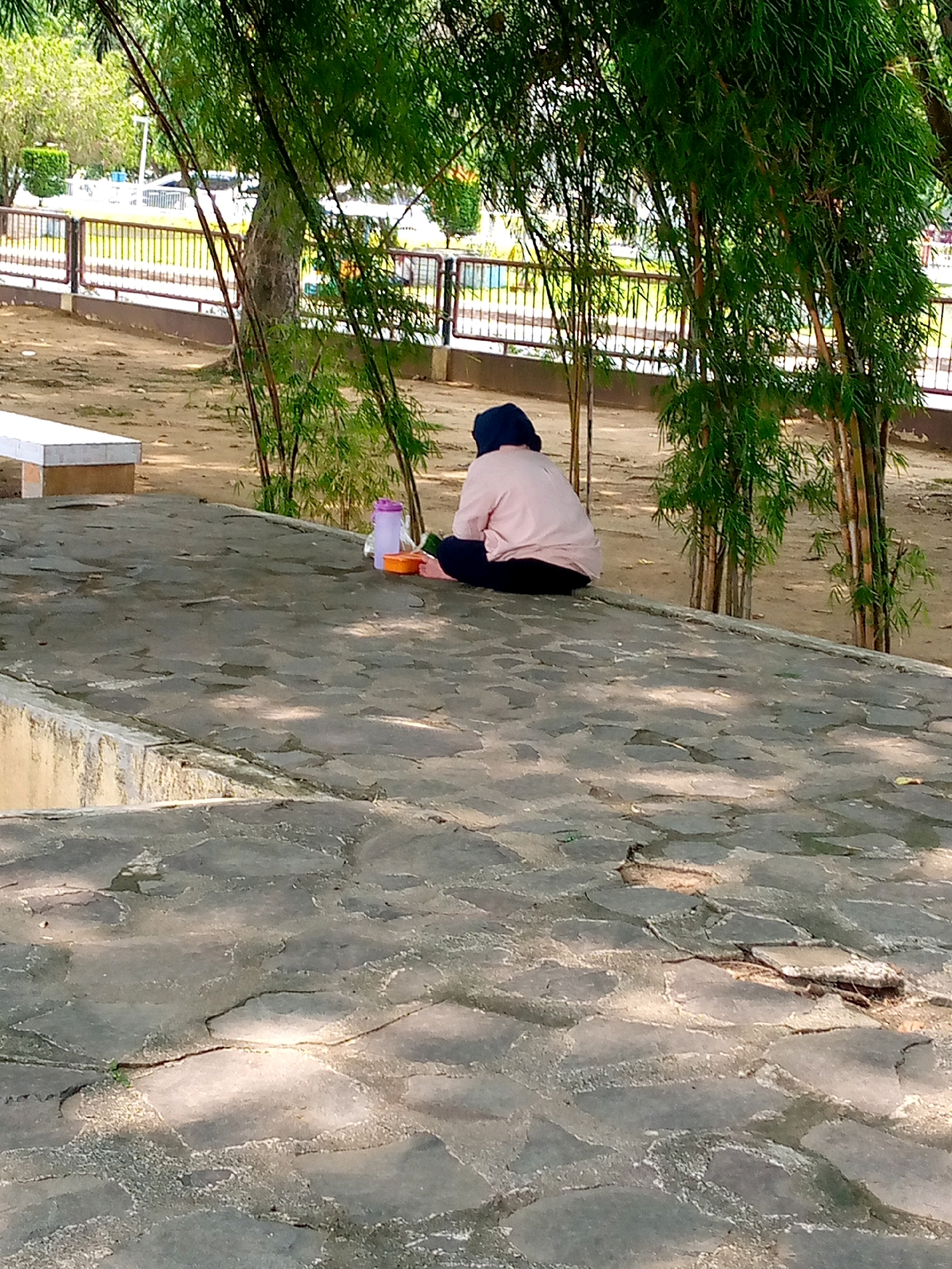 A person wearing a blue headscarf and a pink long-sleeved shirt is sitting on a paved area, facing away from the camera. They are positioned between a light-colored water bottle and an orange container. The paved area is made of irregular grey stones and has visible cracks and some vegetation growing through. Behind the person, bamboo trees with green foliage create a natural backdrop. In the background, a metal fence runs along a path, beyond which is an area with more trees and some blurred structures that suggest a park or public space. The ground around the paved area appears to be dirt or sand, with scattered leaves. A white bench is visible to the left of the paved area. The lighting suggests it is daytime, with shadows cast by the bamboo. The scene is likely located in a park or recreational area in Lhokseumawe, Indonesia, given the typical vegetation and infrastructure often found in such public spaces in the region.