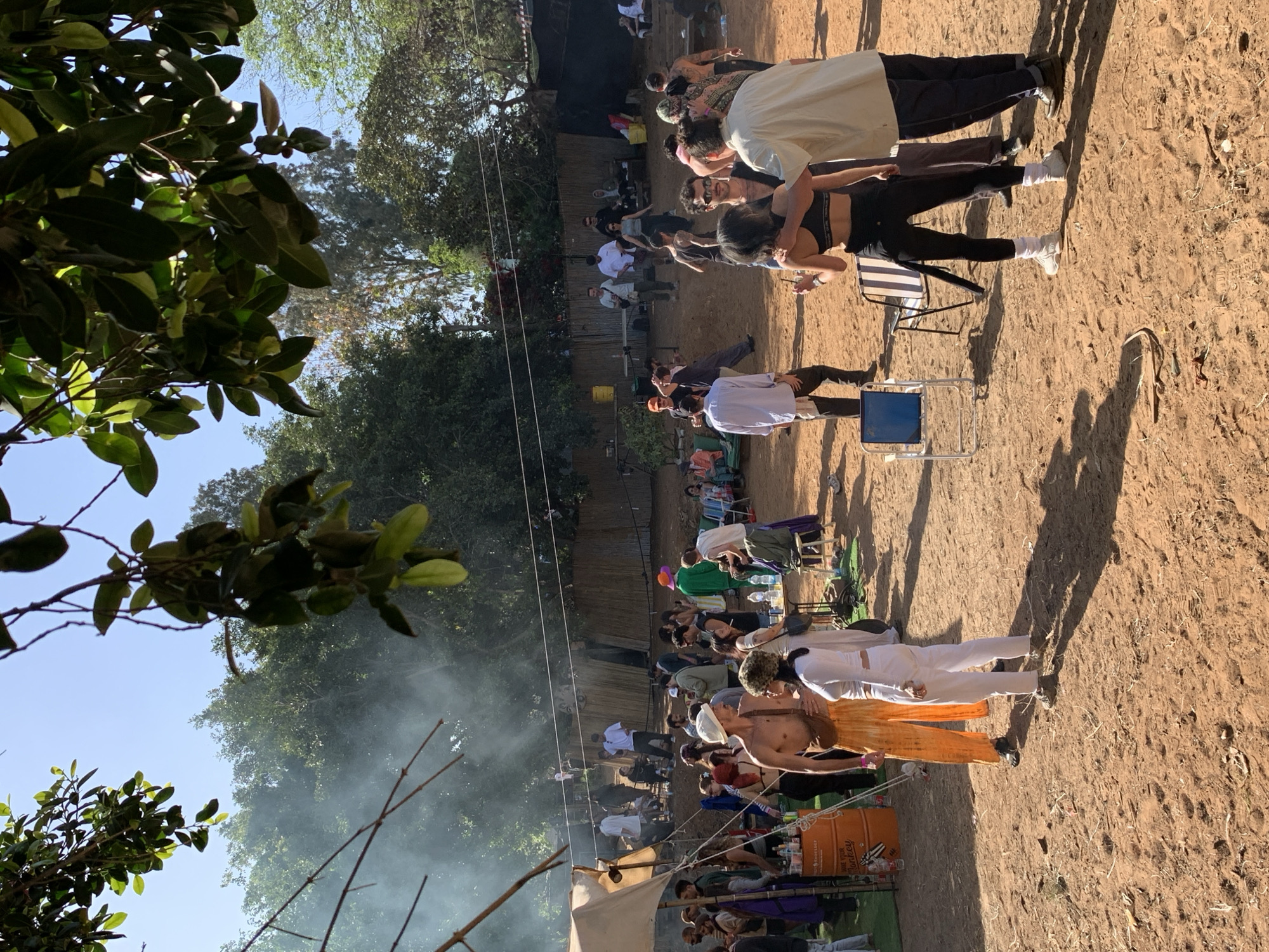 The image shows an outdoor gathering in Gan Soreq, Israel. A large group of people is present, some seated in chairs and others standing. Several people are engaging in what appears to be couples yoga. A food and drinks station appears to be set up near the bottom of the frame, with an orange drum barrel labeled "Woodstock Brewery." Trees and foliage surround the gathering space. The ground is dirt.