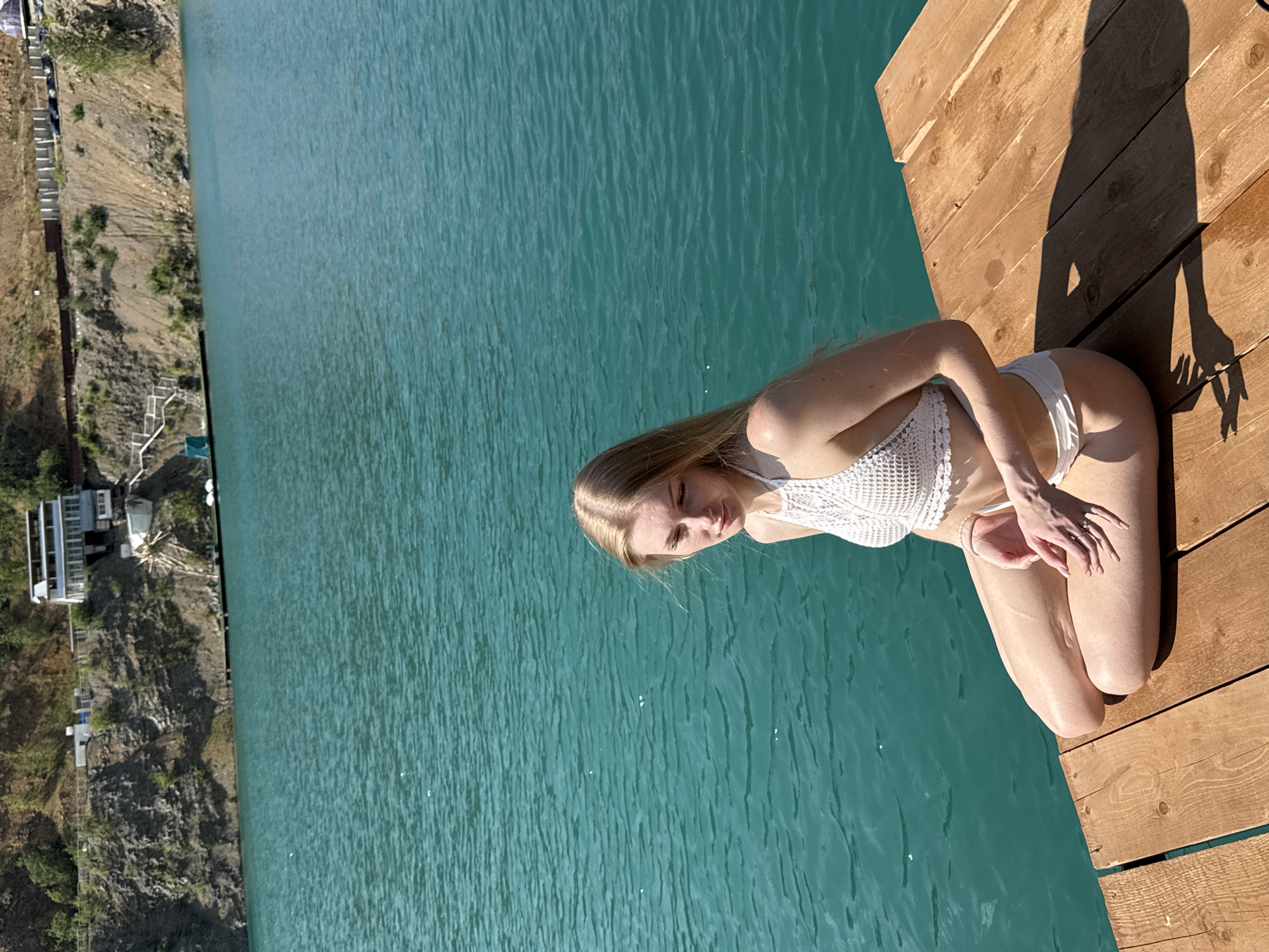 A young woman with blonde hair is sitting on a wooden pier next to a body of turquoise water. She is wearing a white crocheted bikini top and white bikini bottoms. The water is calm, and the pier is made of weathered wooden planks. In the background, there is a steep, rocky slope with some vegetation and a building on top. The sky is clear and blue. The location is identified as Karyer Izvestnyak, Russia.