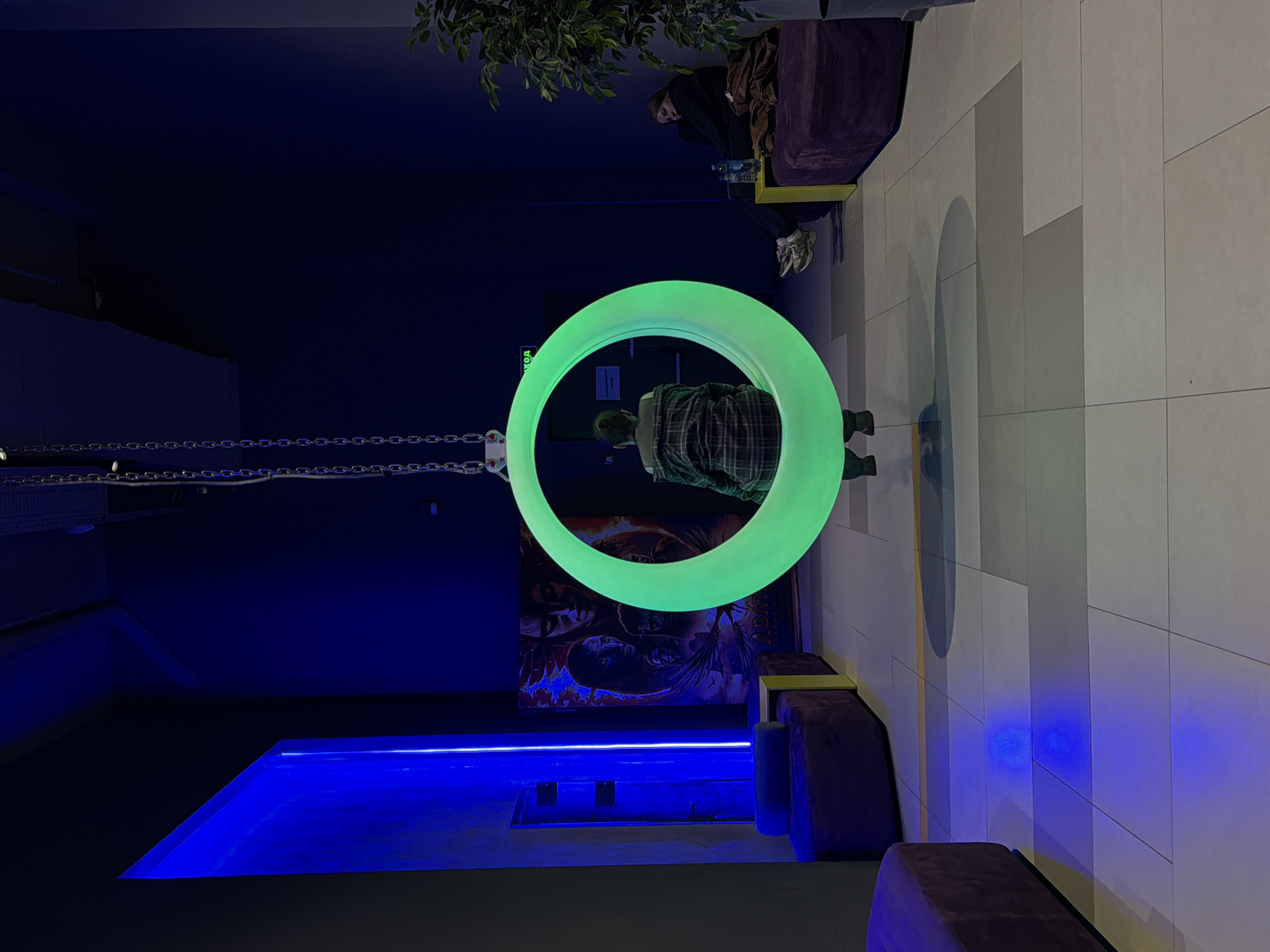 The image shows an indoor space in Penza, Russia, featuring a person standing inside a large, circular, green light fixture suspended by chains from the ceiling. The person is wearing a plaid shirt and faces away from the camera. Behind the light fixture, there's a poster on the wall, and a rectangular pool of bright blue light on the floor. A chain connects the circle to the ceiling on the left side of the image. A person sits in a cushioned chair in the background, partially obscured by a leafy plant. The floor is covered in white tiles.
