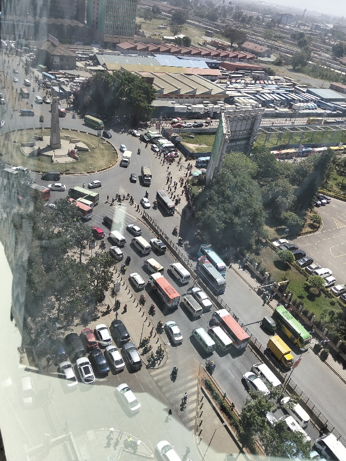 The image provides an aerial, slightly oblique view of a bustling urban intersection in Nairobi, Kenya, specifically depicting the Ambassador Roundabout and its surrounding areas, including parts of the Machakos Country Bus Station and the Nairobi Railway Station vicinity. The scene is illuminated by clear daylight, casting distinct shadows from vehicles and structures, indicative of a mid-day period.
At the center of the foreground is a large circular road, the Ambassador Roundabout, featuring a prominent, light-colored obelisk-like monument on a raised, grass-covered island. Multiple multi-lane roads converge upon and extend from this roundabout. These roads are heavily congested with diverse vehicular traffic, including numerous passenger buses (matatus and larger coaches) in various colors such as red, green, blue, yellow, and white, alongside a multitude of cars, vans, and motorcycles. Many vehicles appear to be stationary or moving slowly.
To the upper right of the roundabout, a large open-air bus terminal, identified as the Machakos Country Bus Station, is visible, densely packed with rows of parked buses, many predominantly blue and white. Adjacent to this terminal are several low-rise industrial-style buildings with multi-colored corrugated roofs. Further in the background, an extensive network of railway tracks and associated infrastructure confirms proximity to the Nairobi Railway Station. On the upper left background, modern high-rise buildings characteristic of a central business district are discernible.
Numerous pedestrians are observed on sidewalks, utilizing designated crosswalks, and gathered in groups near bus stops and the bus station, indicating active movement and waiting. A large, partially visible advertising billboard structure with a green frame stands prominently on the right side of the main road, partially obscured by trees. Green foliage from trees and shrubs lines the roadsides, covers the roundabout island, and forms small patches within the urban environment. The image exhibits reflections on its surface, consistent with being photographed through a window.