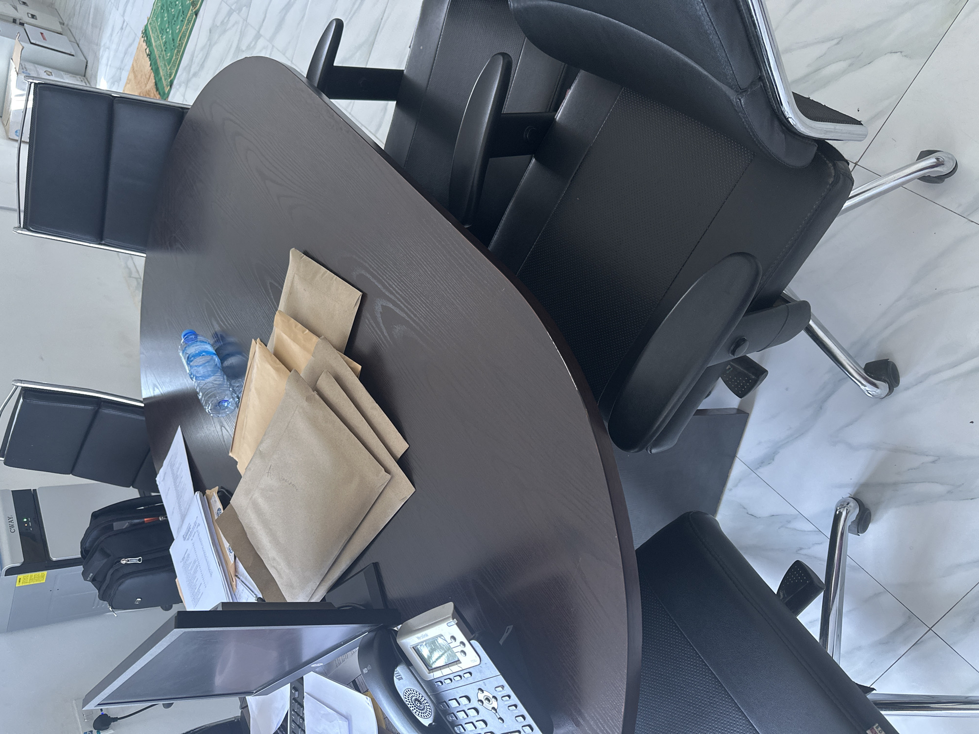 An indoor view captures a dark wood oval conference table with a black leather executive chair visible in the foreground and another to the right. On the table, a stack of manila envelopes and a bottle of water are present. To the left, a computer monitor, keyboard, and a grey office phone are situated. In the background, a black backpack rests near a tall, light-colored machine labeled "CWAY." The environment appears to be an office, with a tiled floor and a portion of a green rug visible in the far background. The overall impression is that of a professional meeting or workspace.