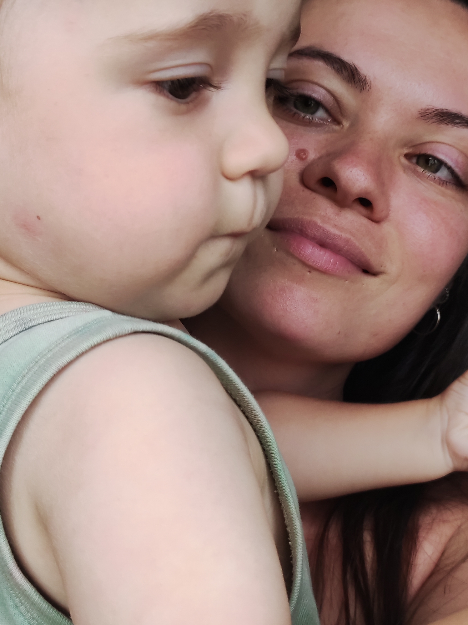 A close-up, indoor, eye-level, vertical shot shows a woman and a young child. The child is on the left, facing away from the camera and to the right, with its cheek pressed against the woman's face. The woman is on the right, looking slightly upwards and to the left. Her right eye and the left side of her face are visible. Both have fair skin. The child is wearing a light green tank top. The woman has dark hair that extends down her right side. The background is out of focus, appearing dark and indistinct. The image appears to be a selfie taken in a home environment. No specific location within Chaikovsky, Russia is identifiable from the image.