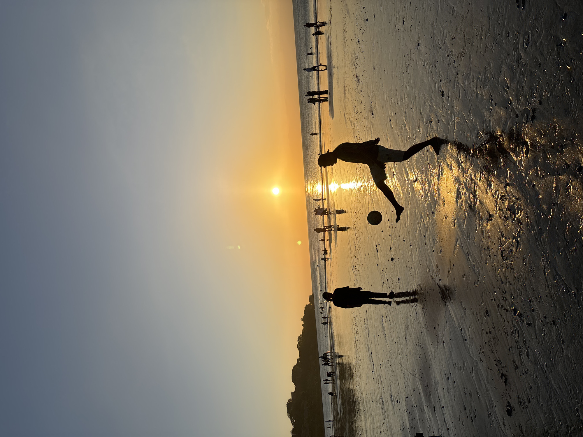 The image captures a sunset scene on a beach in Kuta Selatan, Indonesia. The sun is setting over the ocean, casting a warm, golden glow across the sky and water. The wet sand reflects the sunlight and the silhouettes of people on the beach.
In the foreground, two silhouetted figures are prominent. One, a person in shorts and a t-shirt, is in mid-action, kicking a soccer ball. The other figure, wearing a darker outfit, stands a short distance away, watching.
Scattered across the beach, there are other silhouetted figures, some walking, some standing, and some appear to be engaged in various beach activities. In the background, a hilly coastline is visible, also silhouetted against the bright sky. The ocean stretches out to the horizon, with gentle waves lapping at the shore. The overall atmosphere is peaceful and serene, typical of a beach at dusk.