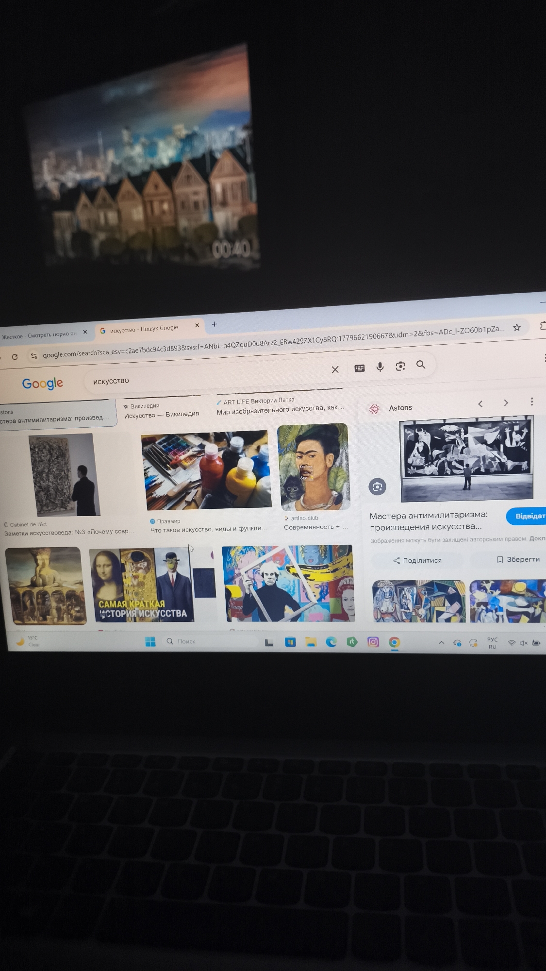 The image shows a laptop screen displaying a Google search results page for "искусство" (art in Russian). The search results include various images of artwork and art-related websites. In the top left corner of the image, there is another screen (likely a TV or monitor) partially visible, displaying an image of a row of houses at nighttime with a timestamp "00:40". The laptop is sitting on a dark surface, possibly a desk or table. The keyboard of the laptop is visible in the bottom part of the frame. The overall lighting is somewhat dim, and the image is taken from an angle looking down at the laptop. The image suggests someone is browsing art-related content online while another image is playing in the background. There is no evidence this image is in Nowy Tomyśl, Poland.