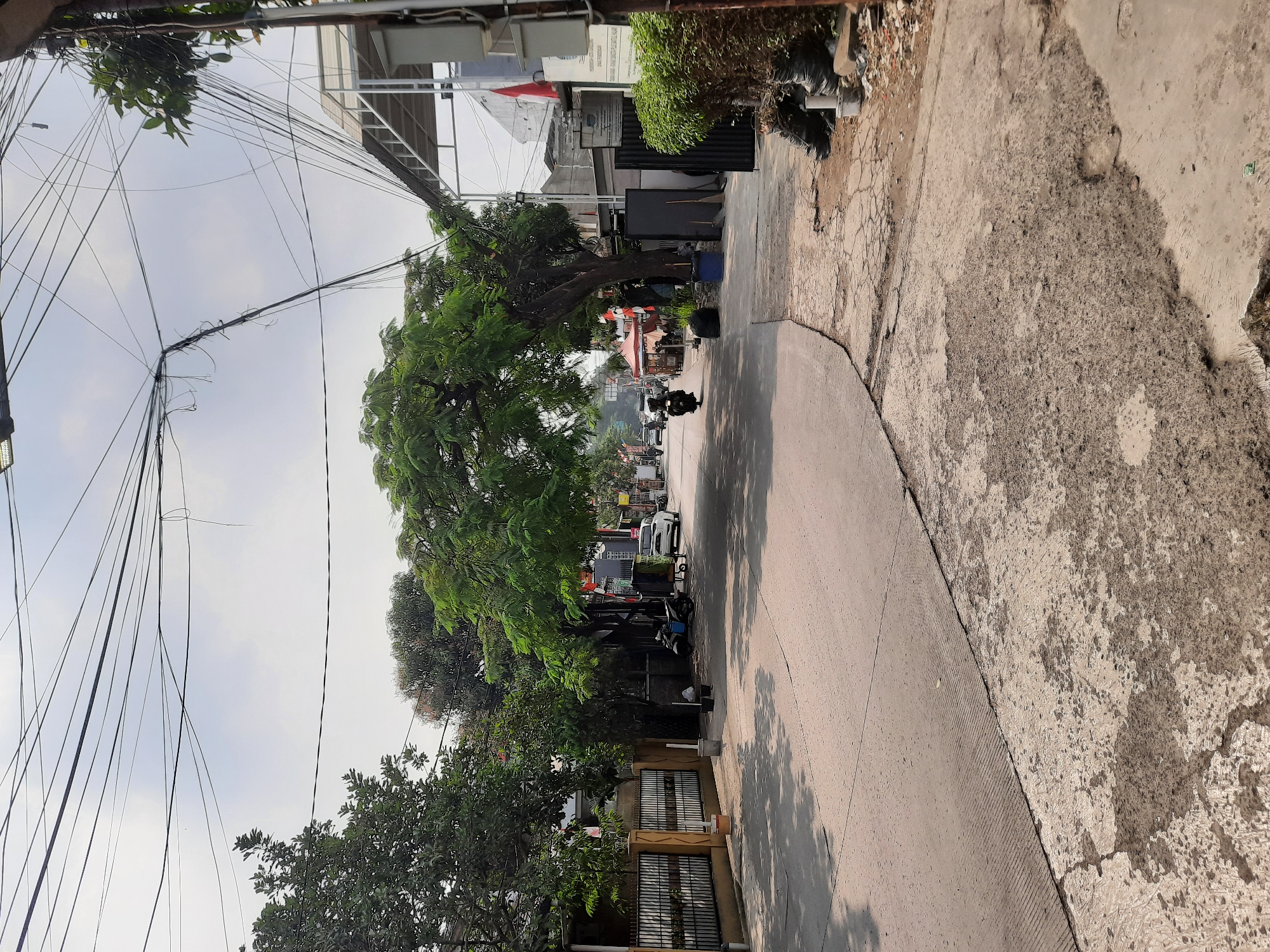 The image shows a residential street in Bandung City, Indonesia, on a partly cloudy day. A paved road curves through the scene, with a broken concrete sidewalk on the right. Lush green trees line the street, with one tree in the center arching over the road. Houses and fences are visible on the left, and buildings with white awnings and flags are on the right. Several motorcycles and a white car are parked or driving on the street. The overall atmosphere is one of a typical urban neighborhood in Indonesia, with a mix of natural elements and developed infrastructure.