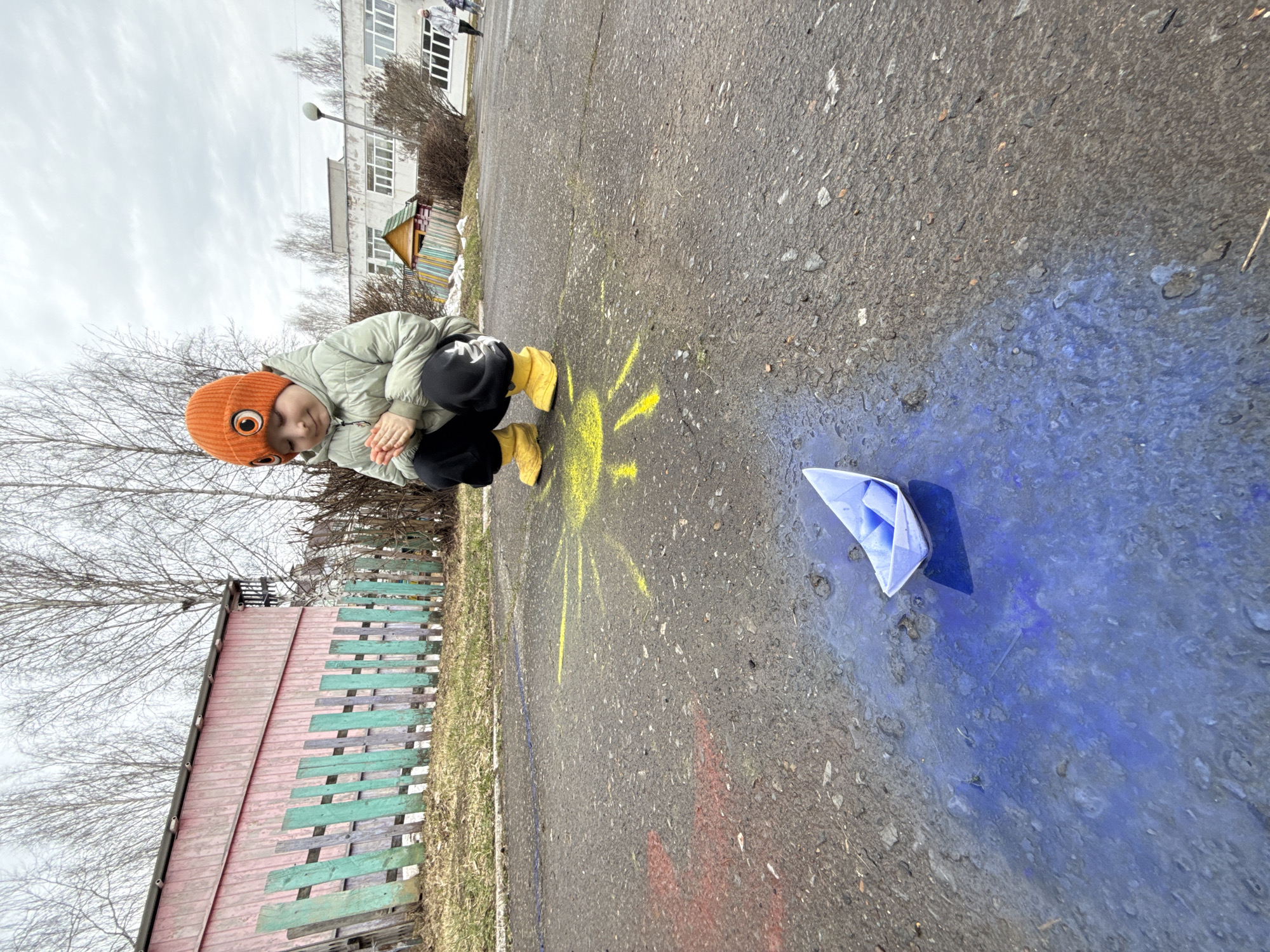 A child wearing an orange hat, a light-colored jacket, black pants, and yellow boots is crouching near a painted sun on an asphalt surface. A blue paper boat is floating in a puddle of blue paint nearby. The background includes a multi-colored wooden fence, bare trees, and a building. The location is the city of Chaikovsky, Russia.