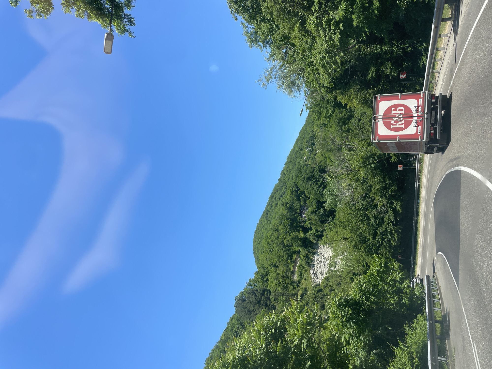 A clear, bright blue sky with faint, wispy clouds is visible above a lush, green mountainous landscape. A winding asphalt road, bordered by metal guardrails, cuts through the scene. On the road, a red truck with a white circular logo featuring red Cyrillic lettering ("К&Б") is driving away from the viewer. The truck has a license plate with Cyrillic characters. The surrounding environment is densely populated with green trees and vegetation, with rocky outcrops visible on the mountainside. A street lamp is mounted on a pole to the right of the road.