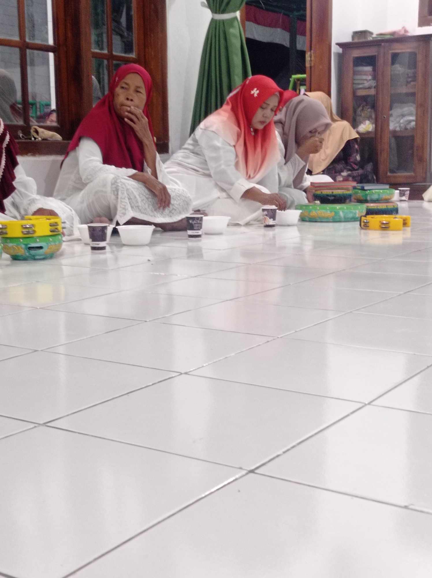 The image shows a group of women wearing hijabs sitting on a tiled floor. The woman on the left is wearing a red hijab and white clothing, with her hand near her mouth. The woman in the center is wearing a pink and orange ombre hijab and white clothing. The woman to her right is wearing a light brown hijab and white clothing, with her hand near her face. In front of them are several bowls, cups, and what appear to be tambourines. In the background, there is a wooden cabinet with glass doors filled with items, and a green curtain. The setting appears to be indoors, possibly a home. The image does not provide enough information to determine the specific location within Maribaya, Indonesia, beyond the general description of an indoor domestic setting.