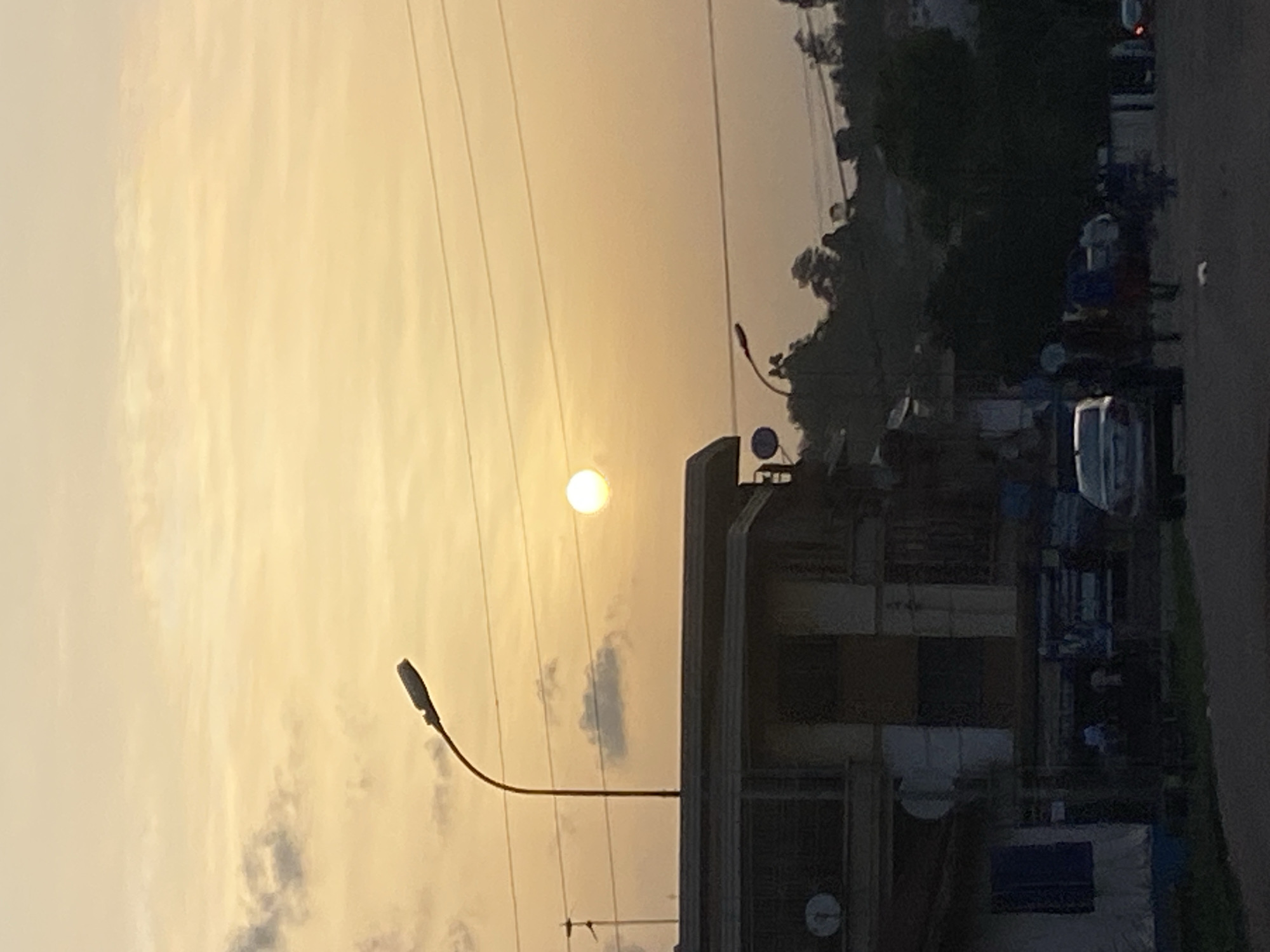 The image shows a view in Abofifia, Nigeria, likely during sunset or sunrise, given the golden hue in the sky. Overhead power lines are visible against the sky. In the middle ground, there is a building with a satellite dish mounted on it. A street lamp extends into the sky. On the right side of the frame, there are parked vehicles and indistinct figures of people, possibly a market area or a roadside gathering. Trees and vegetation are visible in the background. The overall scene appears to be a typical urban landscape in Abofifia during a time of day with low sun angle.