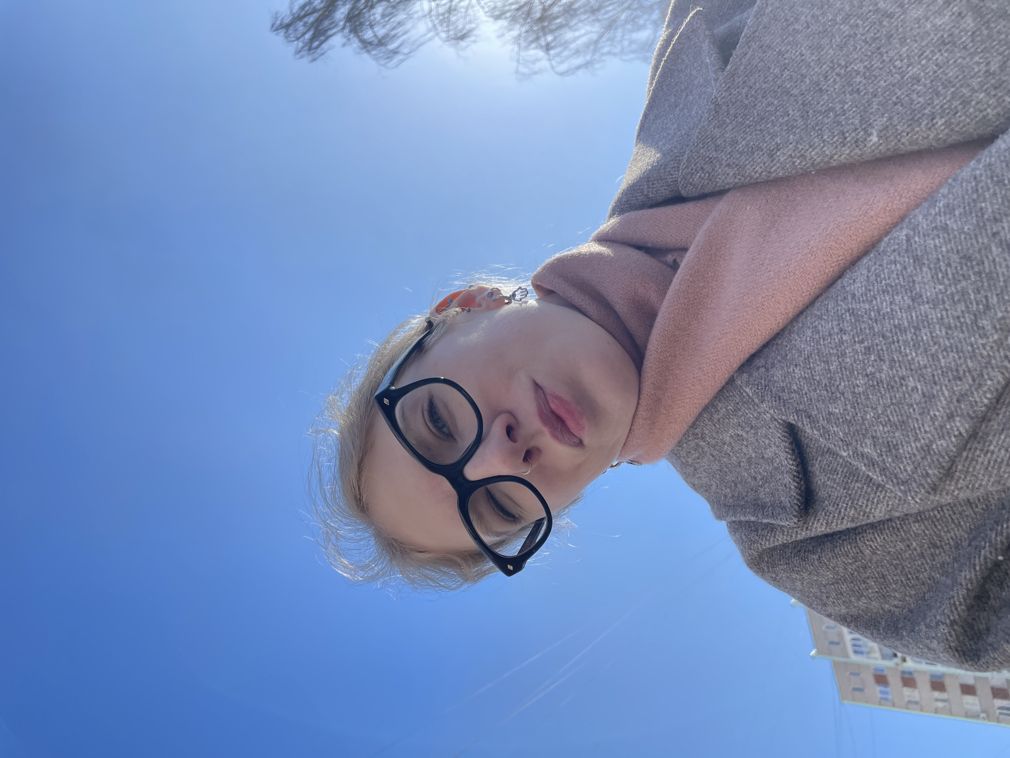 A woman wearing black-framed glasses, a pink scarf, and a gray coat is shown in a low-angle selfie against a clear blue sky. A few bare tree branches are visible in the upper part of the frame, and a multi-story building with light-colored walls is in the lower right. Based on the appearance of the building and the architectural style, it is located in the city of Izhevsk, Russia. The woman's face is angled towards the camera, with a nose ring visible.