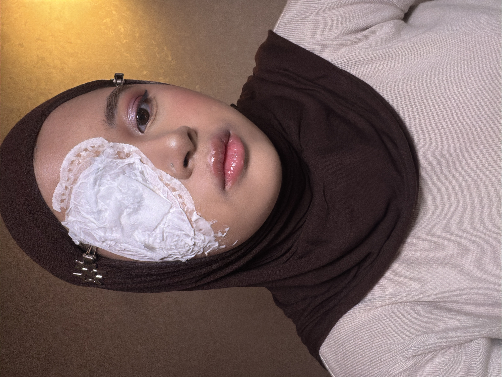 A woman wearing a brown hijab and a light beige sweater lies with her head angled towards the camera. Part of her face is covered in a white substance. Hair clips are visible near her temple. The background is a mottled gold color. The location is East Jakarta, Indonesia.