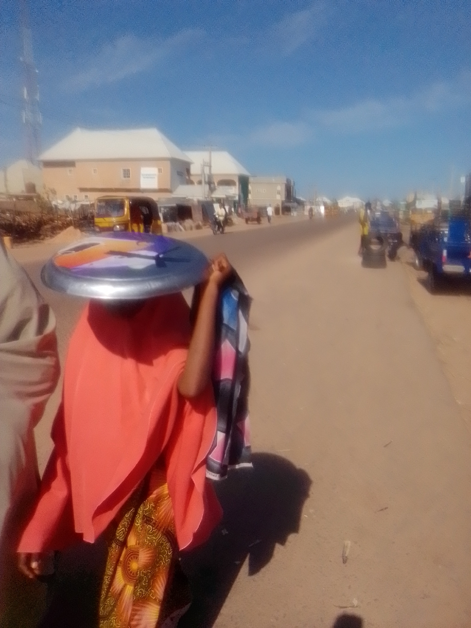 In Pantami, Nigeria, a person covered in an orange cloth is walking along a dusty road. They are balancing a large, round, metallic object on their head, which appears to be a serving tray. The person also holds a bundle of fabric. The road is lined with buildings and a few vehicles, including a yellow bus and a blue truck. The sky is clear and blue, indicating a sunny day. The scene suggests an everyday street view in Pantami, a city located in the Gombe State of Nigeria.