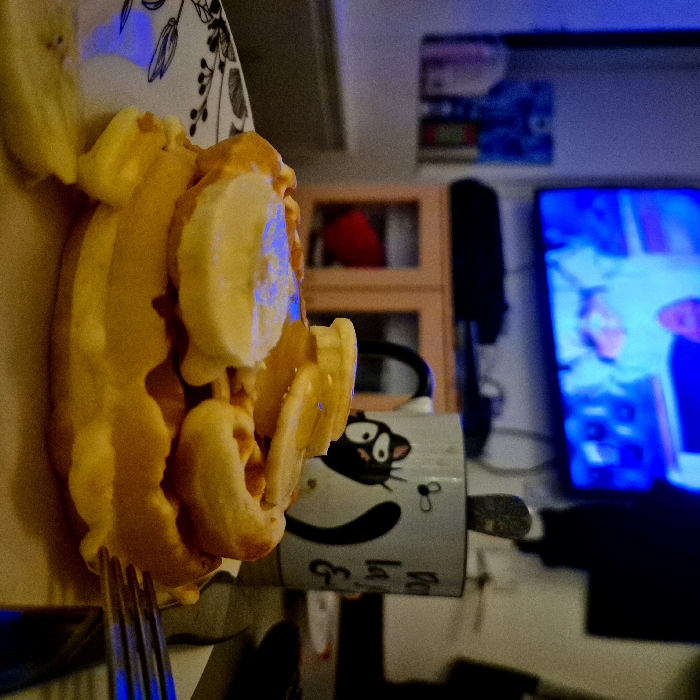 The image depicts an indoor scene within a residence in Strasbourg, France. In the foreground, a round, golden-brown waffle is presented on a white plate with a dark, patterned rim. The waffle is topped with multiple slices of yellow banana and a translucent, glossy drizzle. A silver metal fork is positioned on the right side of the plate, partially obscuring a dark object. To the right of the plate, a white ceramic mug with a black handle features a graphic of a black cat and partial text. A silver spoon is inside the mug. In the midground, behind the food items, a light-colored wooden cabinet with two doors and shelves is visible. The background is dominated by a flat-screen television displaying a brightly illuminated, blue-toned image of an individual in a light-colored garment. The room is dimly lit, with a prominent blue ambient light emanating from the television. A blurry wall-mounted item with various containers is partially visible on the far left.