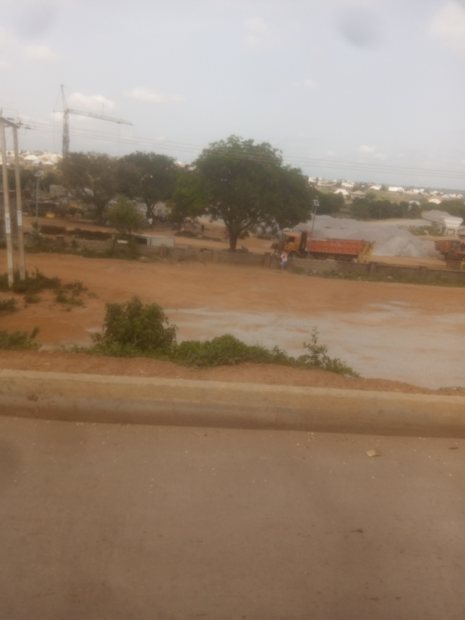 The image depicts a construction site in Sabon Gari Nassarawa, Nigeria. A tall tower crane is visible in the background. In the foreground, a construction area with dirt roads and piles of gravel is apparent. Two orange dump trucks are present, one of which is partially loaded with gravel. A few individuals are scattered around the site, with one person standing near the dump trucks. Trees are sparsely distributed throughout the scene, and a hazy sky suggests overcast conditions. The overall impression is one of ongoing construction and industrial activity.