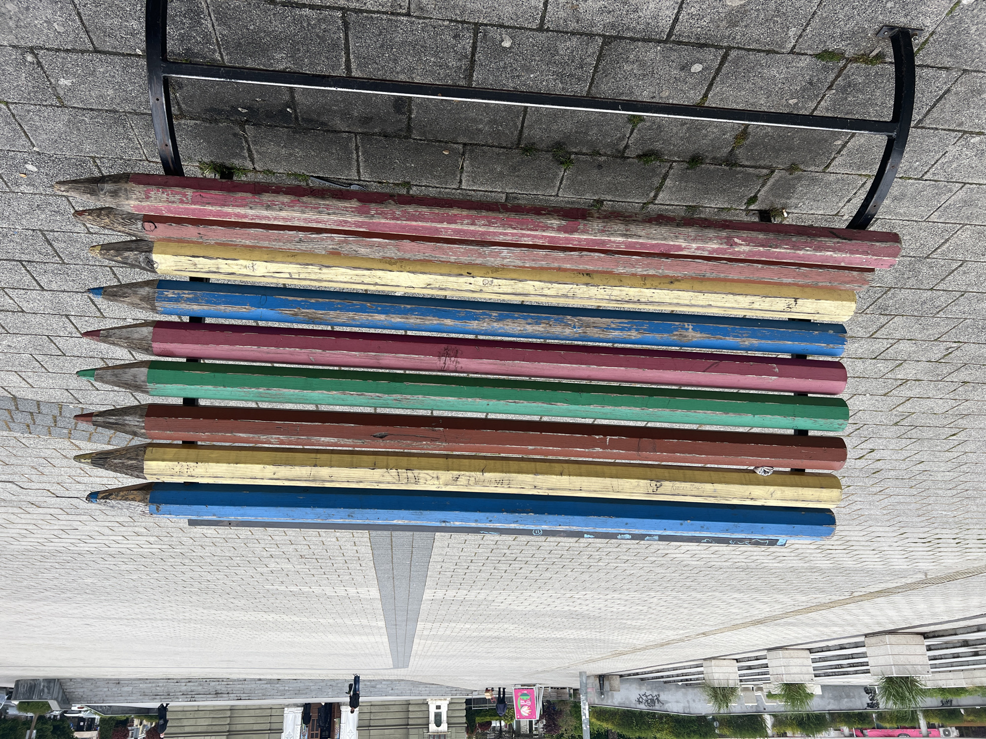 The image captures an artistic park bench designed to resemble a row of large, colorful pencils, situated in an outdoor urban environment, specifically in Burgas, Bulgaria. The bench is composed of ten wooden planks, each shaped like a giant pencil with a sharpened tip visible on the left side, painted in various weathered colors including red, pink, yellow, blue, dark pink, green, brown, and light blue. The paint is chipped and faded in many places, indicating exposure to the elements and frequent use. The bench's support structure is a simple black metal frame visible on the right side.
The bench rests on a gray paved surface, a mix of cobblestones and smoother concrete sections. In the background, beyond the bench, a wide, open public square or promenade unfolds, leading towards distant buildings and some greenery. The ground transitions from a textured paving near the bench to a smoother, lighter-colored surface further back, with a darker gray strip running down the center, creating a sense of depth and perspective.
Several people are visible in the distance, walking or standing, though their faces and details are indistinguishable due to the distance. Some architectural elements, possibly parts of buildings or columns, can be seen in the mid-ground. The weather appears sunny with clear skies, as indicated by the bright, even lighting and the shadows cast on the ground.
Visible text includes faint graffiti or markings on some of the pencil planks. On a yellow plank, "Кристиана" (Kristiana) and "Culo" are faintly discernable. On a blue plank, a circled "⑪" symbol is visible. Further in the background, a pink sign with white, illegible text is present. Graffiti is also visible on some concrete structures in the bottom right of the broader scene. The overall atmosphere is that of a bright, well-maintained public space with a playful, artistic touch provided by the unique bench.