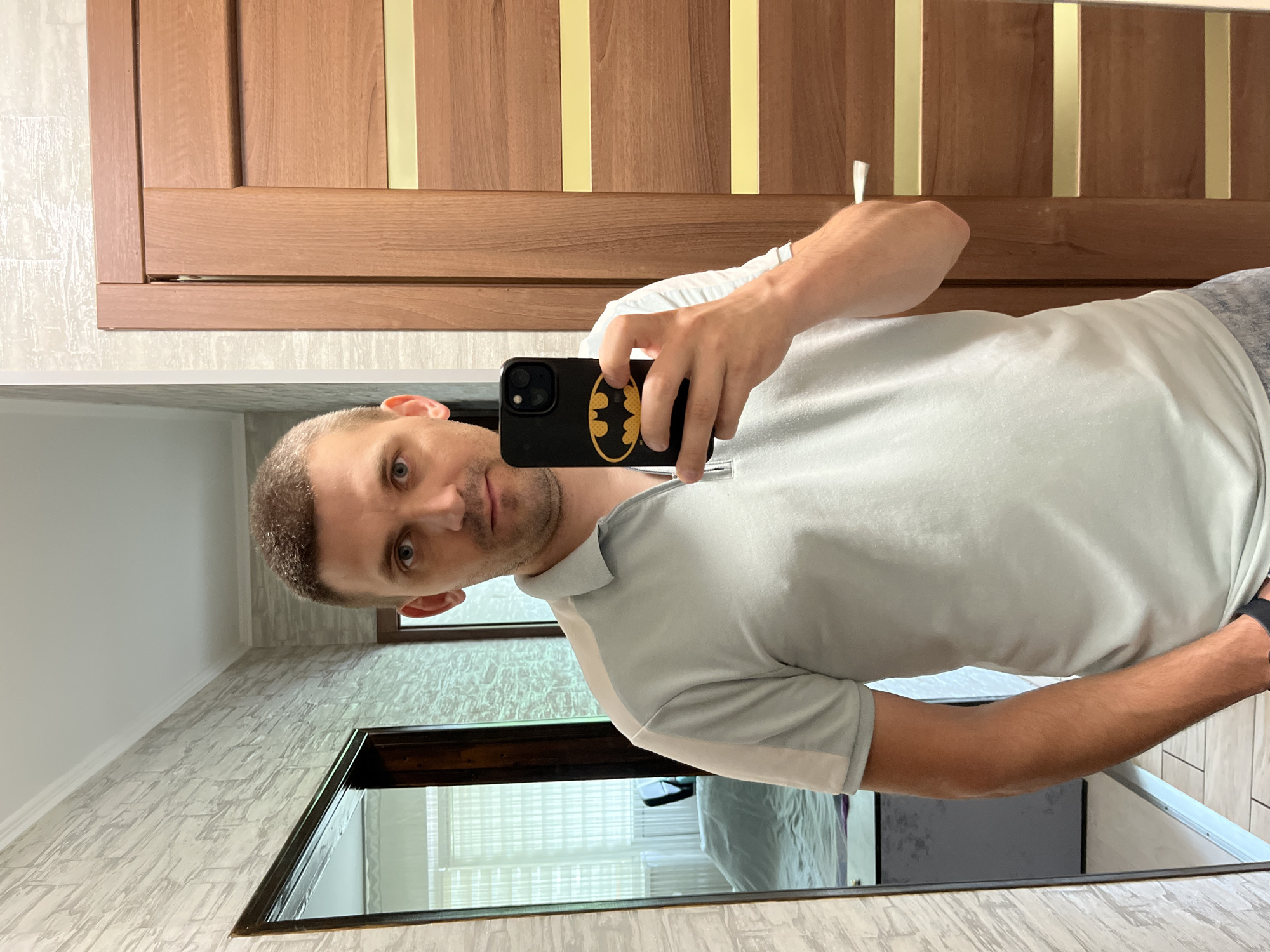 A man with short brown hair and blue eyes, wearing a light blue polo shirt and grey shorts, stands in a room and takes a selfie in a mirror. The man is holding a black smartphone with a Batman logo on its case. To the man's left, a mirror reflects part of a bedroom with a bed and a window with blinds. To the man's right, a wooden door with horizontal yellow stripes is visible. The wall to the man's left has a textured grey wallpaper. The floor is tiled with light-colored square tiles. The image was taken in Kharkiv, Ukraine.