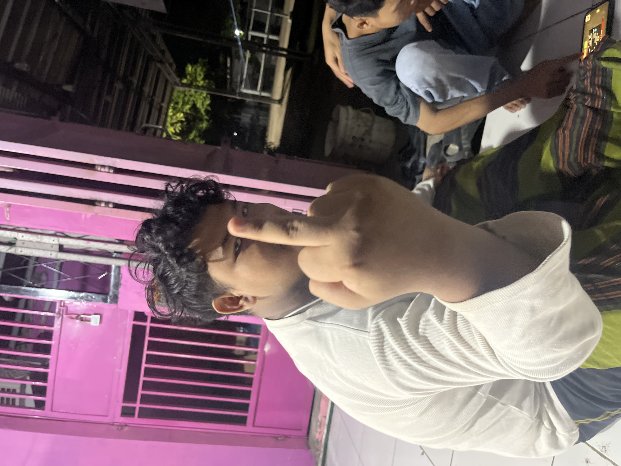 The image is a vertical, medium-shot photograph taken outdoors at night, likely in Laban, Indonesia, given the context. The foreground is dominated by a young person, facing the camera with their right hand raised, displaying their middle finger. They have dark, curly hair and are wearing a white, long-sleeved shirt. Behind this person, to the right, another young person is visible, crouched and looking down at a smartphone, wearing a dark t-shirt and light blue pants. The background features a bright pink metal gate with a padlock, typical of Indonesian residences, and a wooden structure overhead. The ground appears to be tiled. The overall atmosphere suggests a casual, informal gathering at dusk or night.