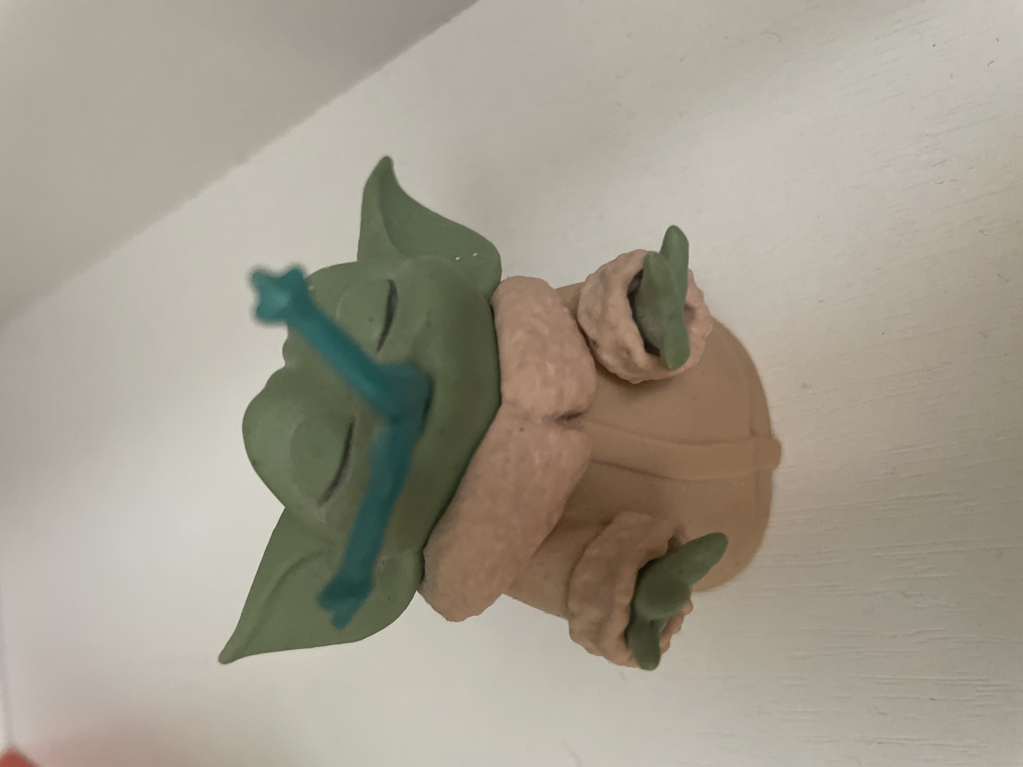 A small, green figurine resembling the character Grogu from Star Wars is positioned on a white, textured surface. The figurine is wearing a tan robe with a pink, furry trim. A teal, stick-like object is placed across the figurine's eyes. The figurine's eyes are closed, and its arms are extended forward. The background is a plain, white wall. The image was taken in Moscow, Russia.
