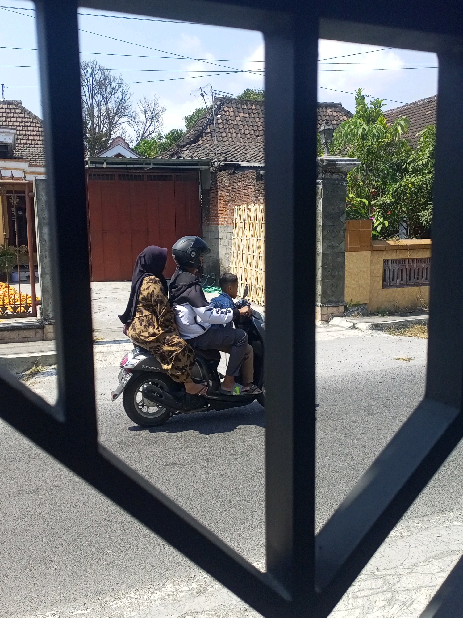 A medium shot shows three people on a scooter traveling on a paved road in the city of Ngasem, Indonesia. The scooter is black, and the two rear passengers, a woman and a child, are on the back. The woman is wearing a dark headscarf and a patterned brown dress. The child is wearing a blue shirt. The driver is wearing a black helmet. In the background, there is a brick wall, a large red gate, trees, and a house with a tiled roof. The scene is viewed through the bars of a black metal fence in the foreground. The sky is blue with scattered clouds.