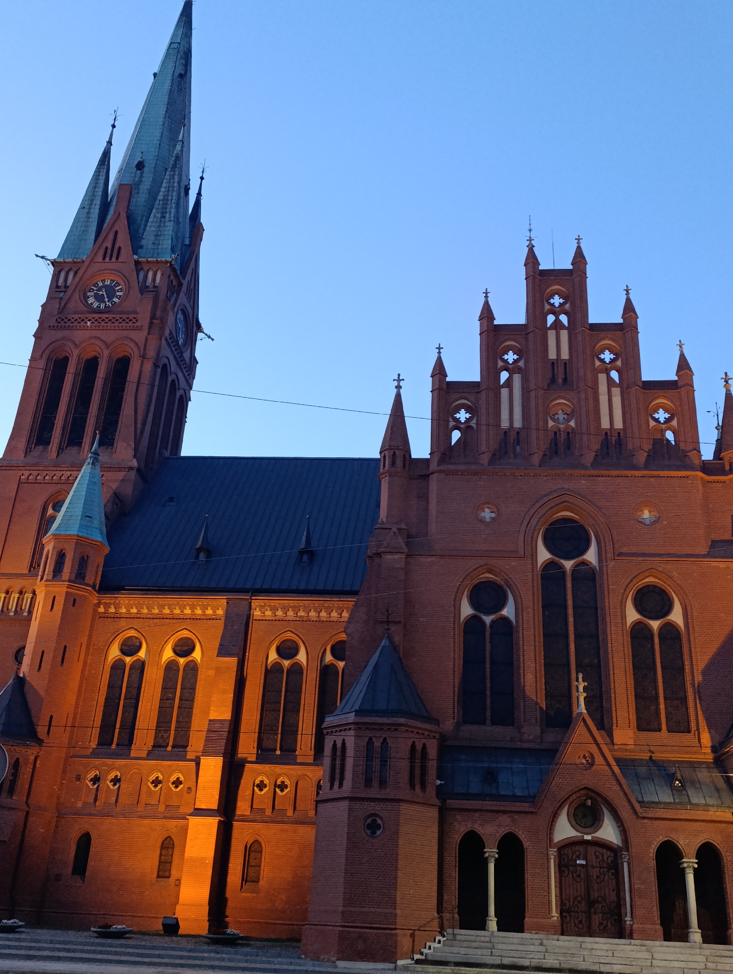 The image shows the exterior of St. James' Church in Toruń, Poland, at dusk. The Gothic Revival church is constructed primarily of red brick. The prominent feature is a tall, slender bell tower with a green spire, topped with a cross. A clock face is visible on the tower. Adjacent to the tower is the main body of the church, characterized by pointed arch windows and decorative brickwork. The facade has multiple gables and smaller spires, also capped with crosses. The entrance features arched doorways with ornate double doors, flanked by columns and steps. The building is illuminated by warm, orange artificial lights, casting shadows and highlighting the architectural details. The sky above is a clear, pale blue, typical of twilight. No people are visible in the image.