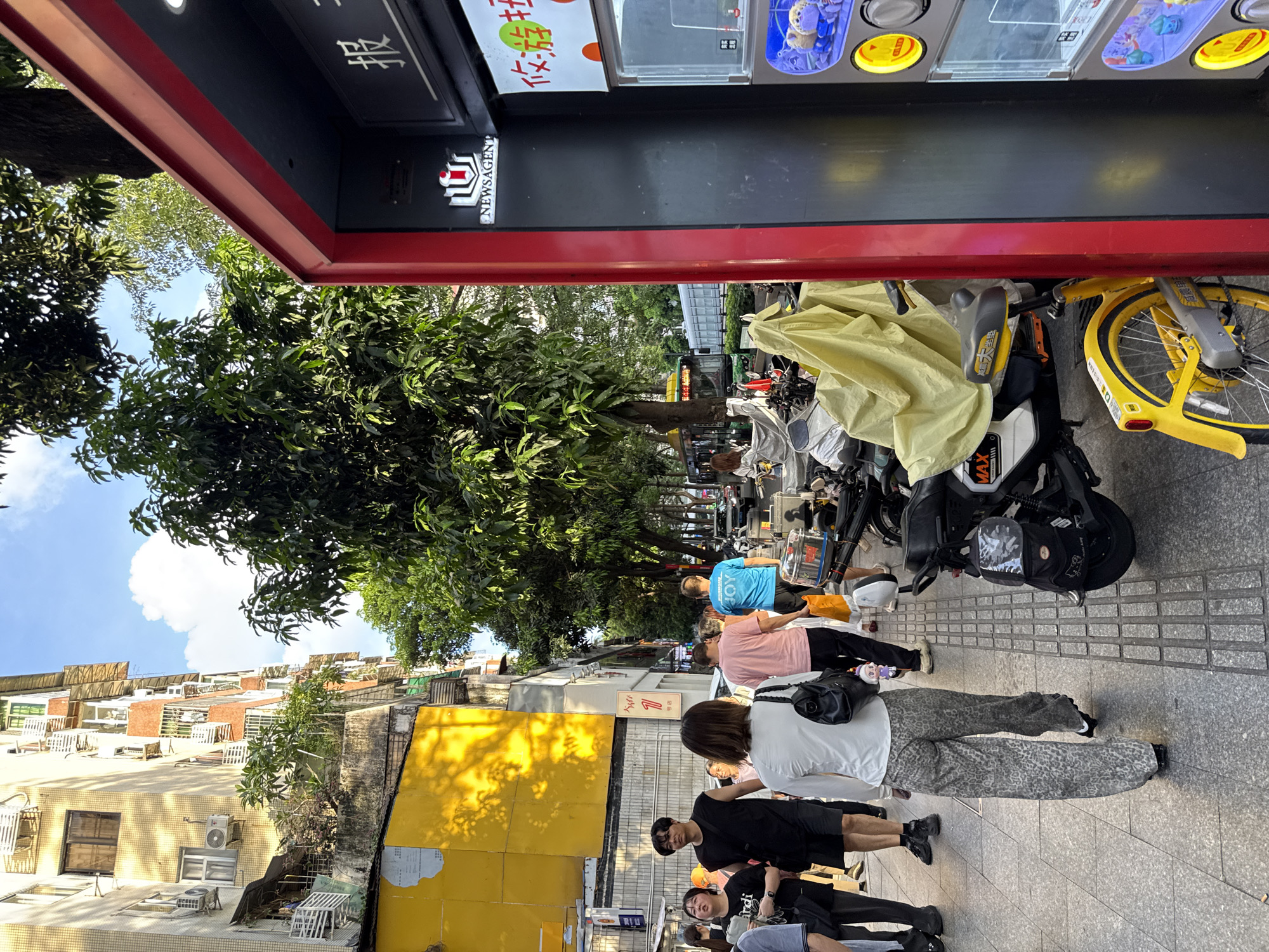 On a sunny day in Haizhu District, China, pedestrians walk along a sidewalk next to a street lined with trees and buildings. In the foreground, a red and black vending machine stands on the right side of the image, displaying items and Chinese characters. Several parked electric scooters, some covered with yellow tarps, are visible on the sidewalk. A group of people are walking away from the camera towards the left. One woman wears a white long-sleeved shirt and grey leopard-print pants, carrying a black bag. A man in a black sleeveless shirt and shorts walks ahead of her. Another woman in a pink t-shirt and a man in a blue t-shirt with the word "JOY" printed on it are also visible in the group. The buildings on the left are multi-story structures with visible air conditioning units and balconies. The street is paved with gray square tiles and is lined with trees. In the background, traffic, including cars and a bus, can be seen on the street. The sky is blue with scattered clouds.