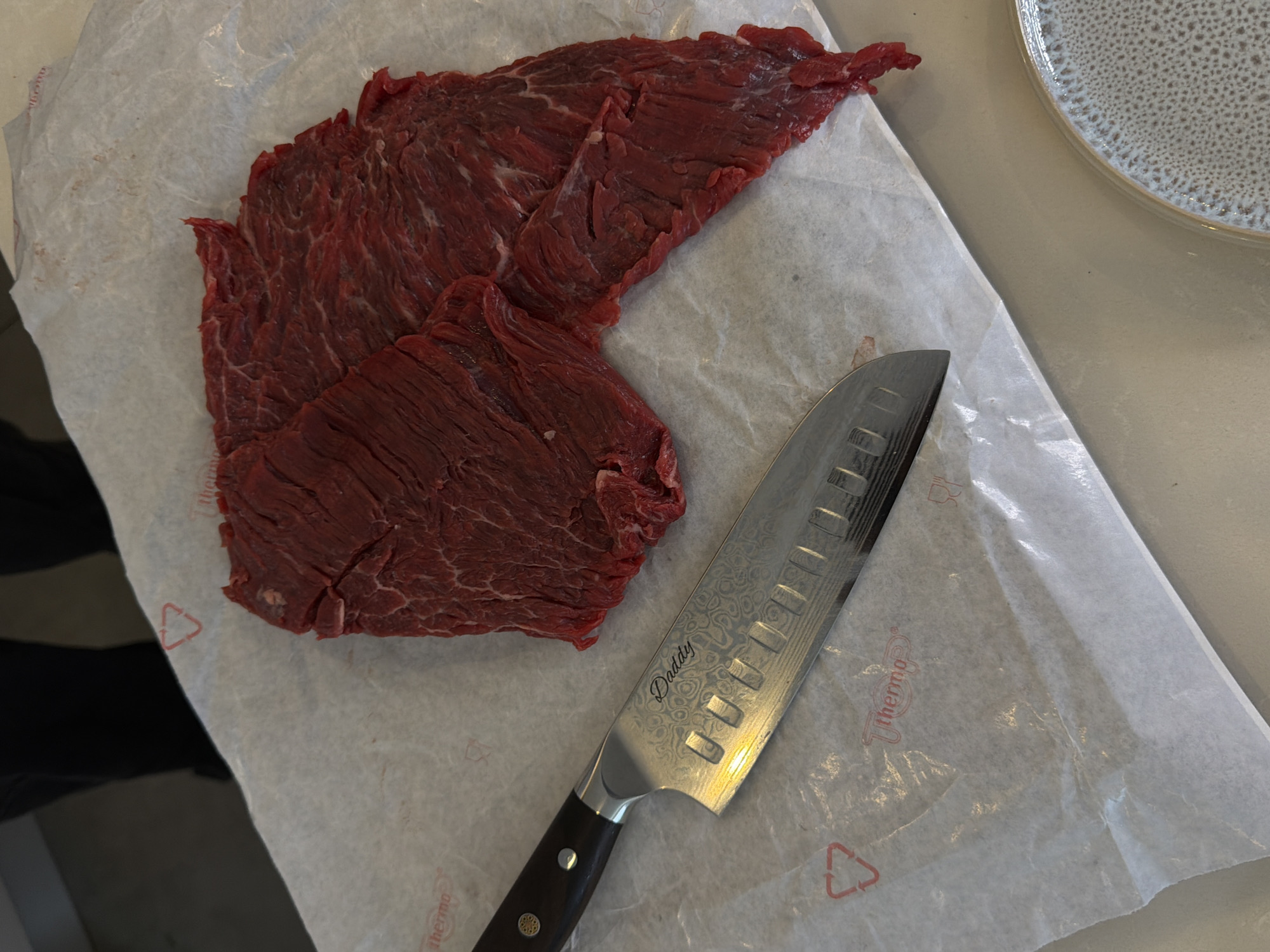 A top-down shot captures raw, thinly sliced red meat, likely beef, laid out on white, somewhat crinkled baking or butcher paper. The meat, rich red with visible muscle fibers, occupies the left and central-left portions of the frame. To the right of the meat, a gleaming chef's knife or santoku knife rests on the paper, pointing towards the top right corner.
The knife features a distinctive patterned blade, possibly Damascus steel or an etched design, and has Granton-style indentations along its edge. Prominently engraved on the blade in a cursive font is the word "Daddy." The knife's handle appears to be made of dark wood or composite material, secured with silver-colored rivets and a decorative element at its base.
The paper underneath the meat and knife bears faint red printed details, including a triangular recycling symbol and the word "Thermo," which is visible in multiple spots. In the upper right corner, a segment of a round, ceramic plate with a textured, speckled off-white glaze is visible. The setting suggests an indoor environment, likely a kitchen or food preparation area, with the raw ingredients and tools ready for cooking. No specific time of day or emotional cues are discernible, and the focus is on the still life arrangement of food and utensils. The image captures a moment of food preparation in what could be a domestic setting.