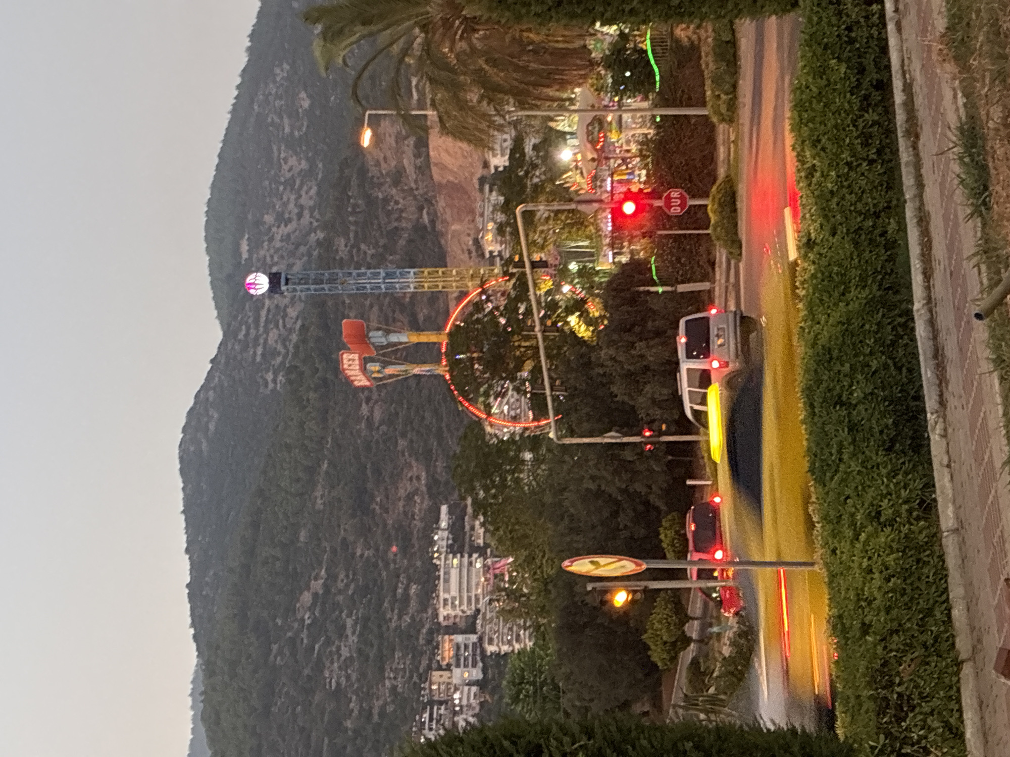 The image shows a street scene in Alanya, Turkey, with an amusement park in the background. The amusement park features several rides, including a tall tower with a lit sphere at the top, a "Ranger" ride with a swinging arm, and a circular ride with red lights. In the foreground, there is a street with cars, traffic lights, and a "DUR" (stop) sign. The sky is overcast, and the overall lighting suggests it is either dawn or dusk. The landscape behind the amusement park is hilly and covered with trees, with buildings visible on the slopes. Green hedges line the foreground and a paved area. One car in the foreground has motion blur, indicating it is moving. A traffic light shows red.