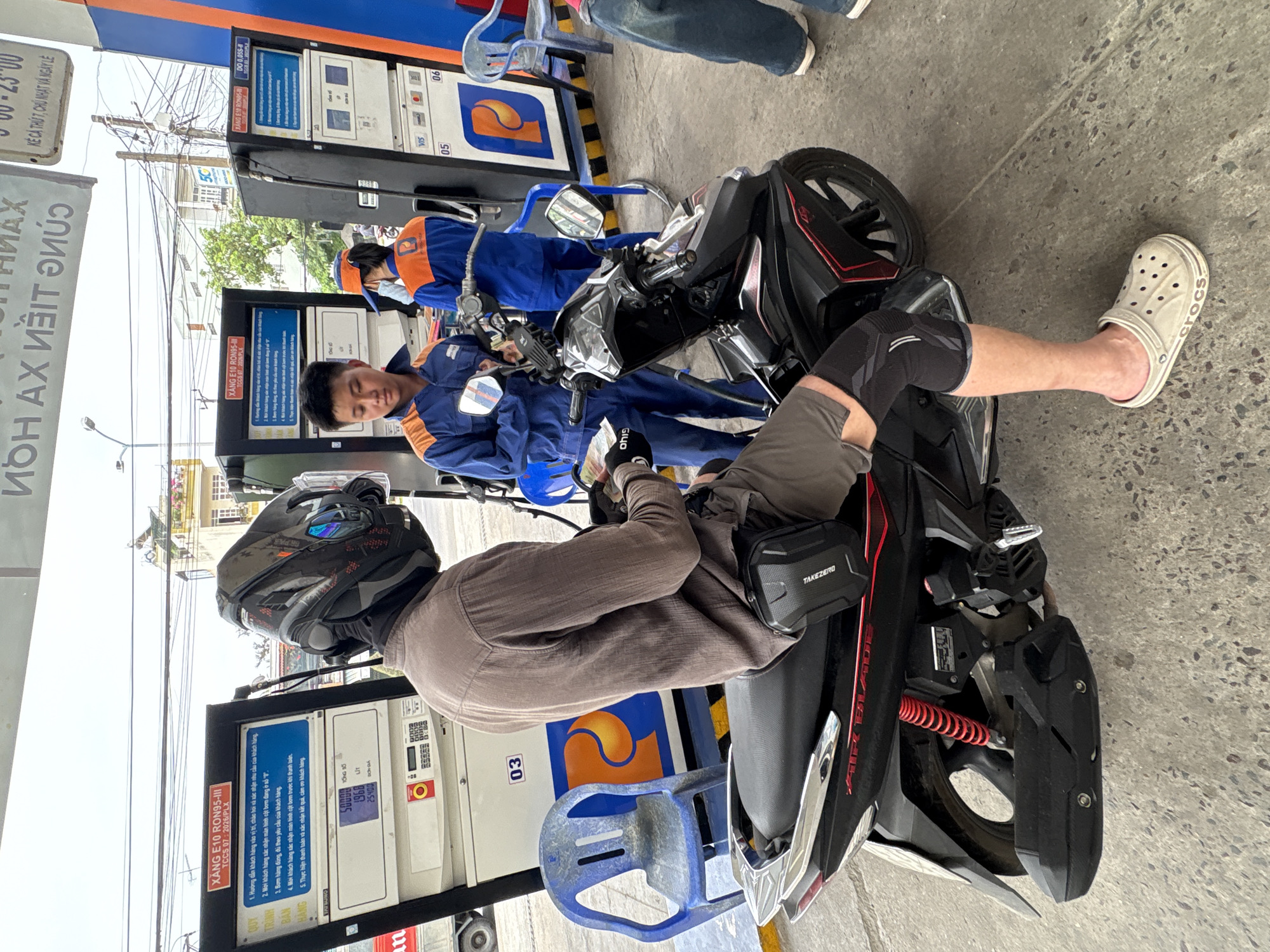 The image, taken at what appears to be a petrol station in Bac Nha Trang Ward, Vietnam, shows two men and a motorcycle. One man is seated on the motorcycle, wearing a helmet, a brown shirt, and a knee brace, with a "TAKEZERO" bag attached to the side of the motorcycle. He is also wearing a white "Crocs" sandal. Another man, wearing a blue uniform with orange accents, is standing next to the motorcycle, apparently attending to it. A second person in a matching uniform is seen in the background. The motorcycle is a black "Air Blade" model. The petrol station has multiple pumps with signage in Vietnamese, with pumps labeled "03," "05," and "06." Details such as petrol grade, price per liter (1968 VND), and total price (25400 VND) are visible on the pumps. A blue plastic chair is located next to the pump, and overhead power lines are visible in the background.