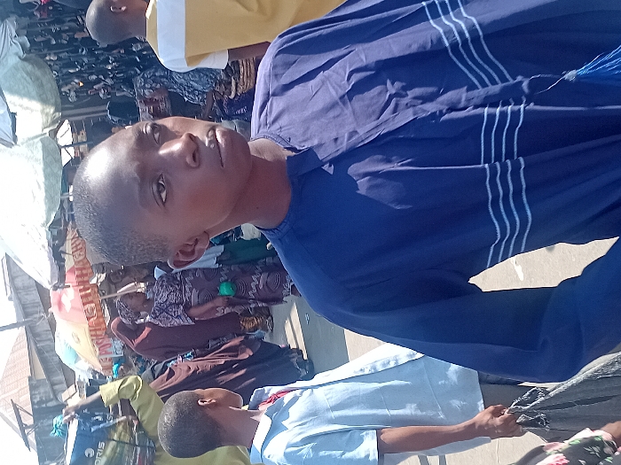 A young man in a blue shirt with white stripes stands near a bustling marketplace. People and stalls fill the background, partially obscured by parasols. One person wears a light yellow shirt, another wears a school uniform. The photo is taken at an angle. The lighting appears to be bright, suggesting an outdoor setting.