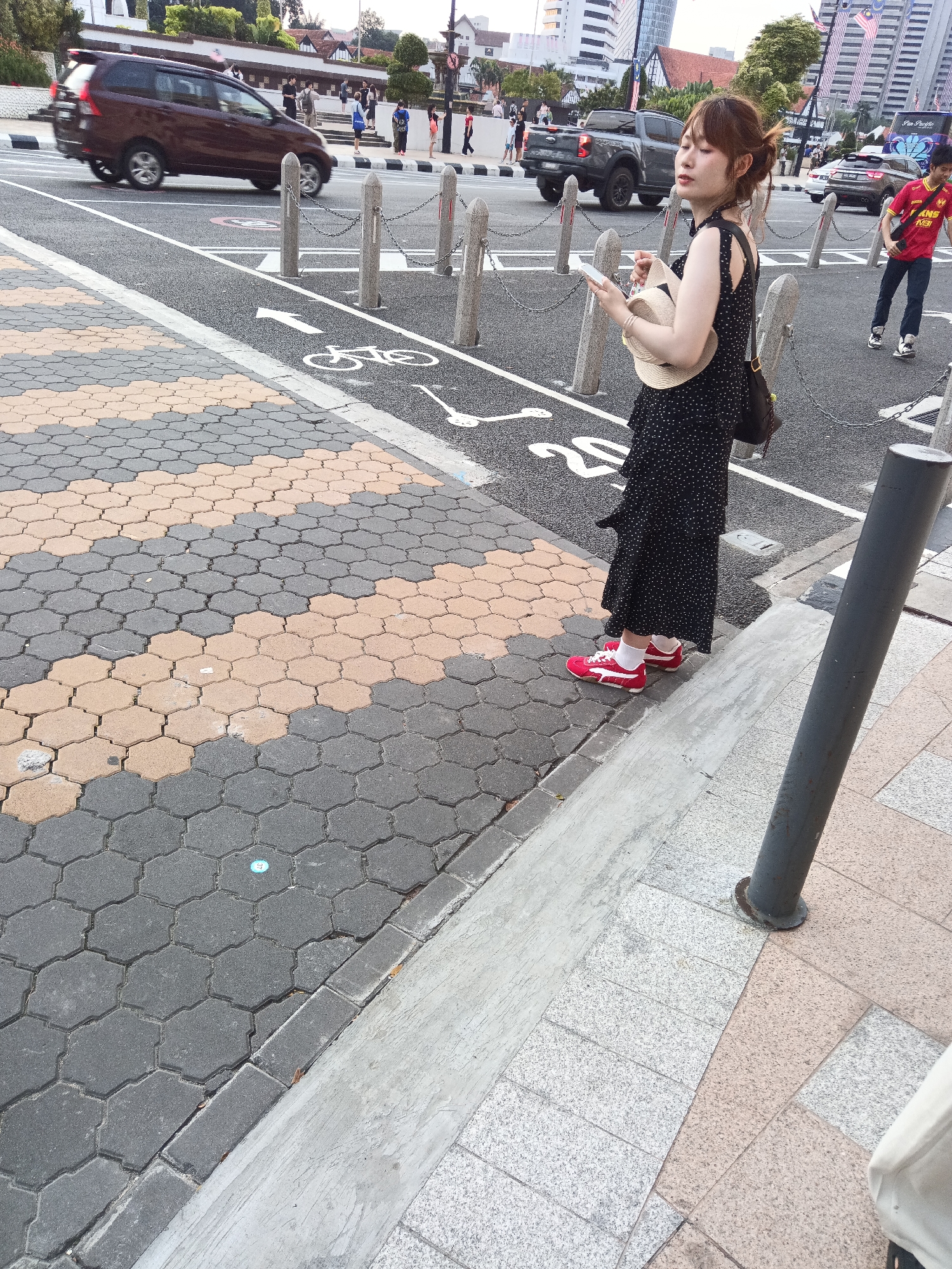 An outdoor medium shot captures a young East Asian woman standing on a street in Kuala Lumpur, Malaysia, near the Merdeka Square area. She is dressed in a black polka-dot dress, white socks, and red sneakers. The woman holds a straw hat in her left hand and a smartphone in her right. Her attention is directed downward towards the phone.
The environment features a distinctive pavement of hexagonal tiles, alternating between grey and a light brown hue. To the right, a grey metal pole stands vertically, positioned on a sidewalk paved with grey and pink speckled granite tiles. In the background, a brown van drives past on the asphalt road, with a bicycle lane marked on the road surface. Beyond the road, there are bollards with chains, a pickup truck, and a few pedestrians. The overall scene suggests a public urban setting during daylight.