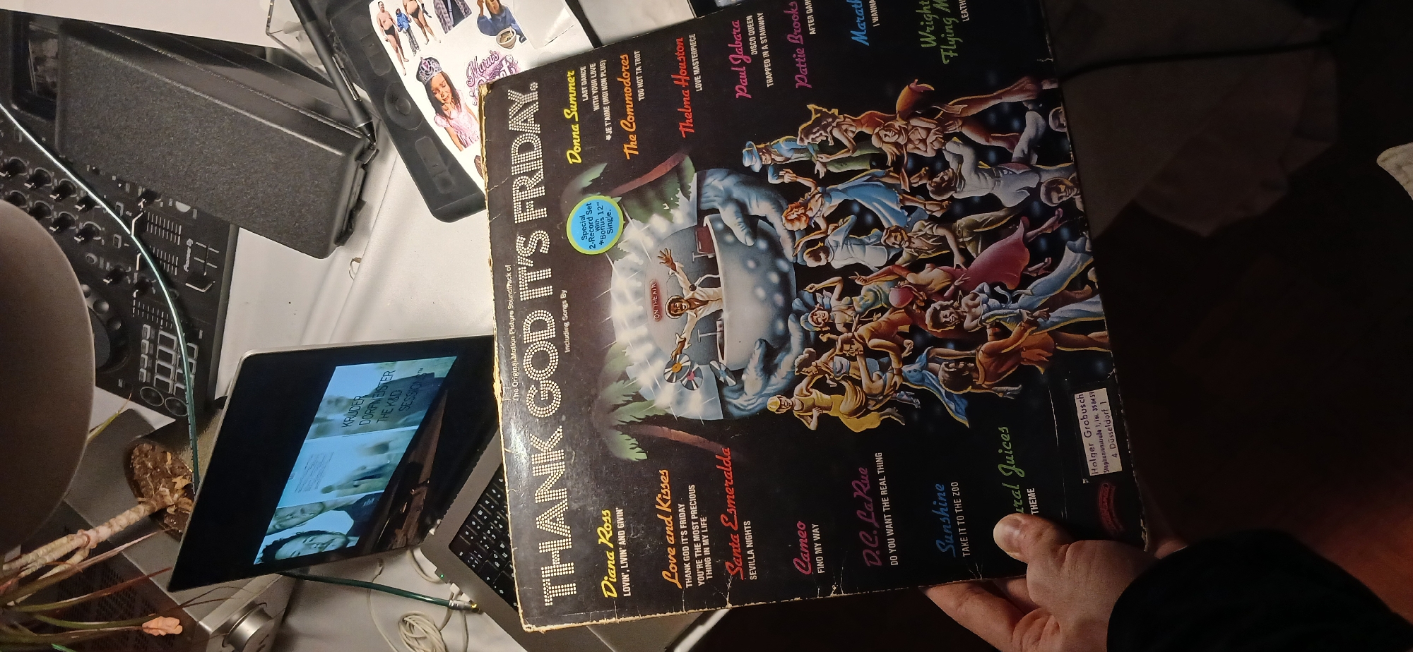 The image shows a vinyl record cover for "Thank God It's Friday", the original motion picture soundtrack, held in a person's hand. The record cover is displayed against a cluttered background including a laptop screen showing the album art for "Kruder & Dorfmeister - The K&D Sessions", a DJ mixer, and other miscellaneous objects. The environment appears to be an indoor space. The location in the city of Unknown, Unknown, is not discernible from the given information.