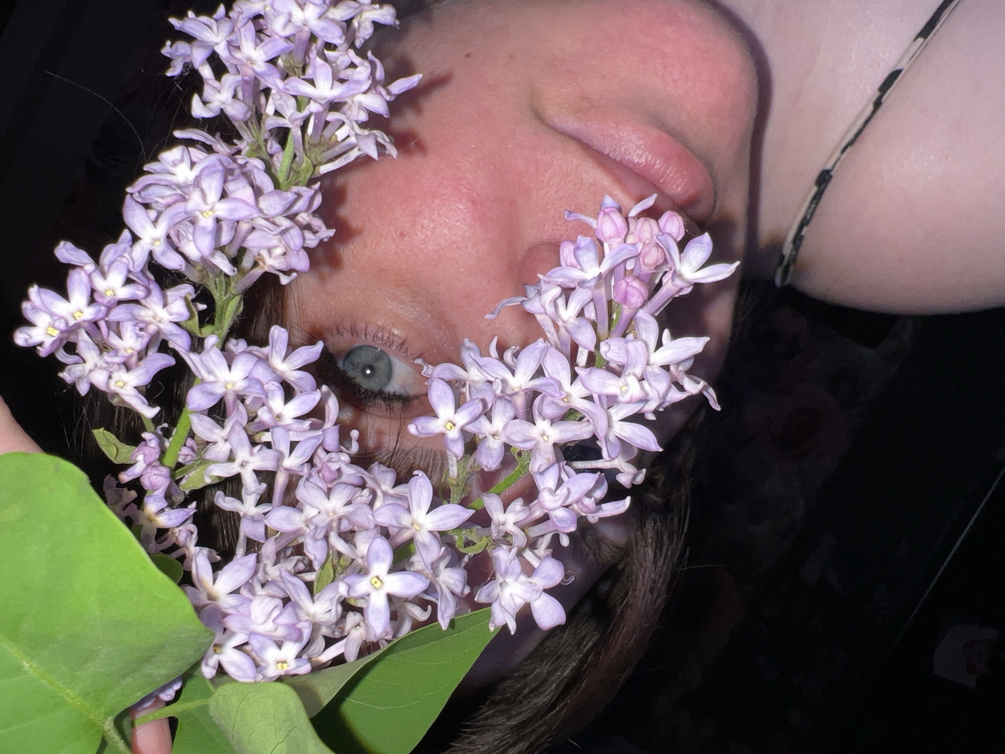 A close-up, rotated view shows a person holding a sprig of purple lilac flowers up to their face. The person is fair-skinned, with one eye visible and light colored. Dark brown hair is seen in the lower part of the frame. A thin strap suggests a garment worn by the person. The background is mostly dark and unclear. There is no city or location in this image.