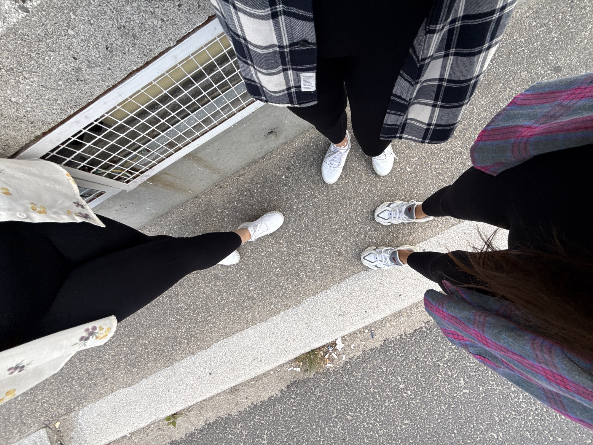The image shows the legs and feet of three individuals standing on a sidewalk next to a paved area. One person is wearing black leggings, a white and floral patterned shirt, and white sneakers. Another person is also wearing black leggings and white sneakers. The third person is wearing black leggings, a black and white plaid shirt, and white sneakers. They are all standing near a building with a grated window opening. The ground is a mix of asphalt and concrete, with some debris on the sidewalk. The setting appears to be an outdoor urban or suburban environment. Based on the information provided, this image was taken in the city of Arefu, Romania.