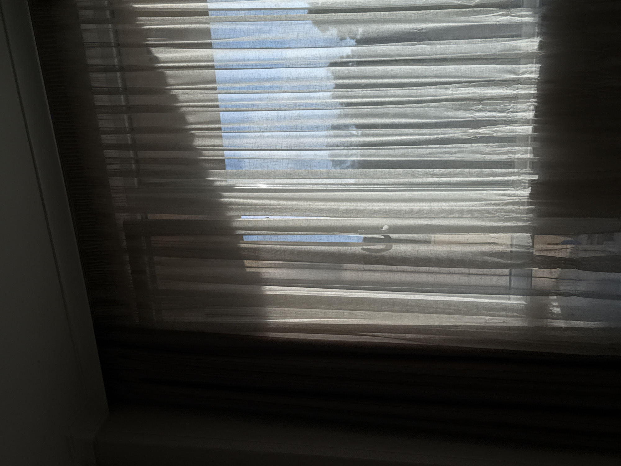 The image shows a window with closed blinds, partially obscuring a view of the sky. The blinds are a light color, possibly beige or white, and appear to be made of a fabric or textured material. The slats are horizontal and create alternating bands of light and shadow. Sunlight is filtering through the gaps in the blinds, casting shadows across the surface. The sky visible through the gaps is light blue. The window frame and wall around the window are visible, and are painted white. The location is unknown, but the prompt indicates the location is in the city of Varna, Bulgaria.